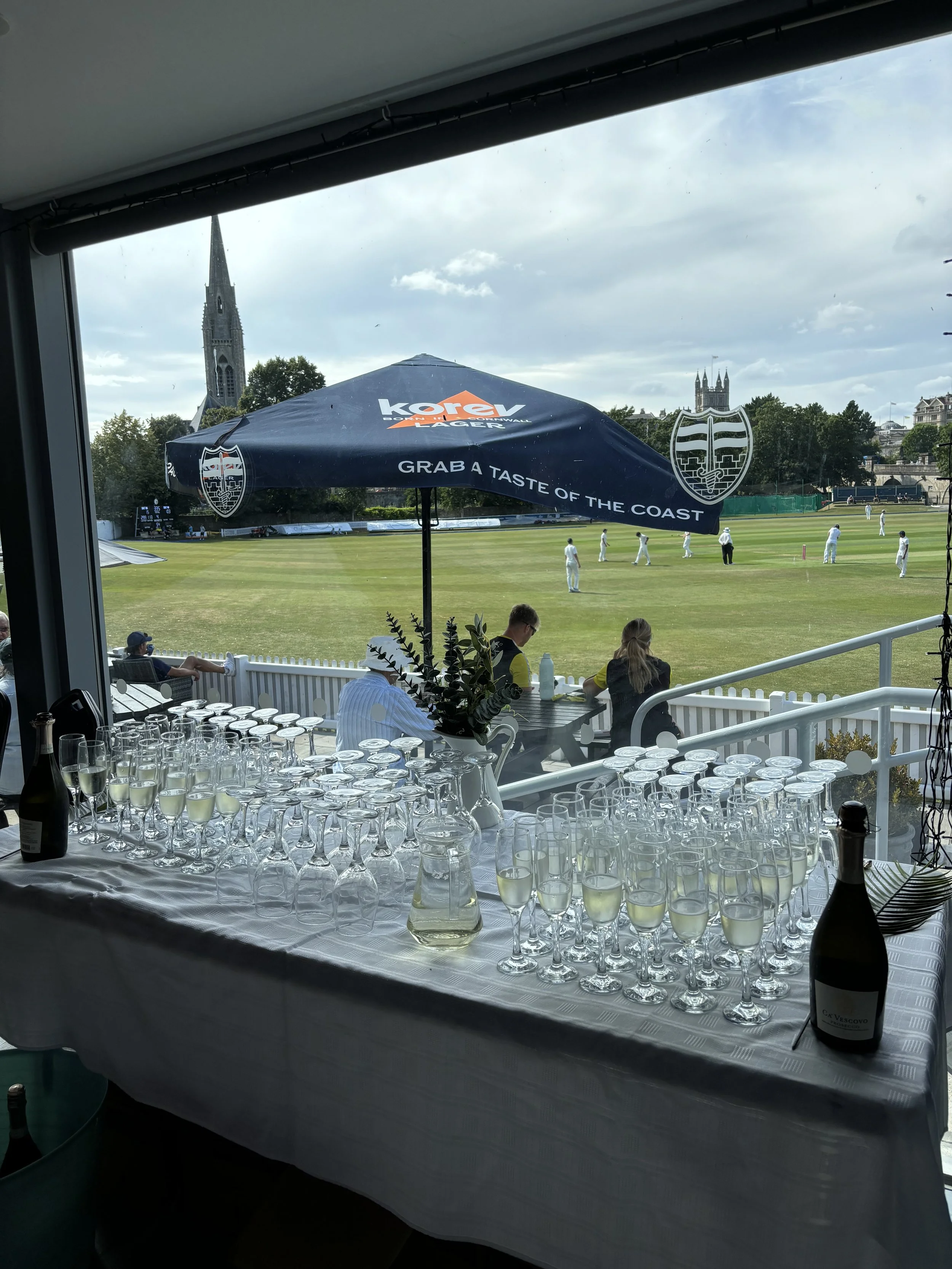 Table with glasses of white wine and bottles, overlooking a cricket match on a lush green field with players in white uniforms and historical buildings in the background.