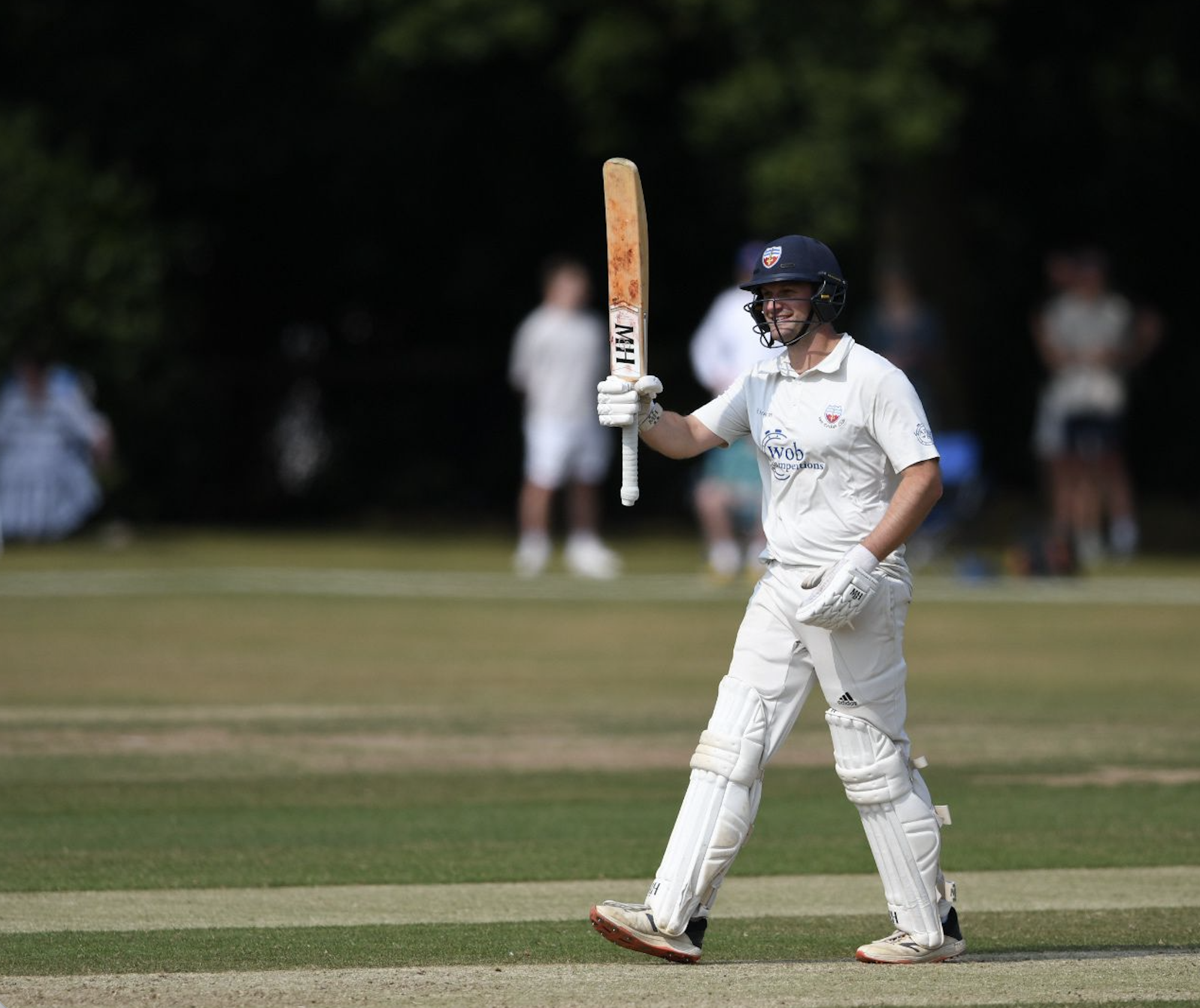 Cricket player in white uniform, wearing a helmet, smiling and holding a cricket bat, on a cricket field.
