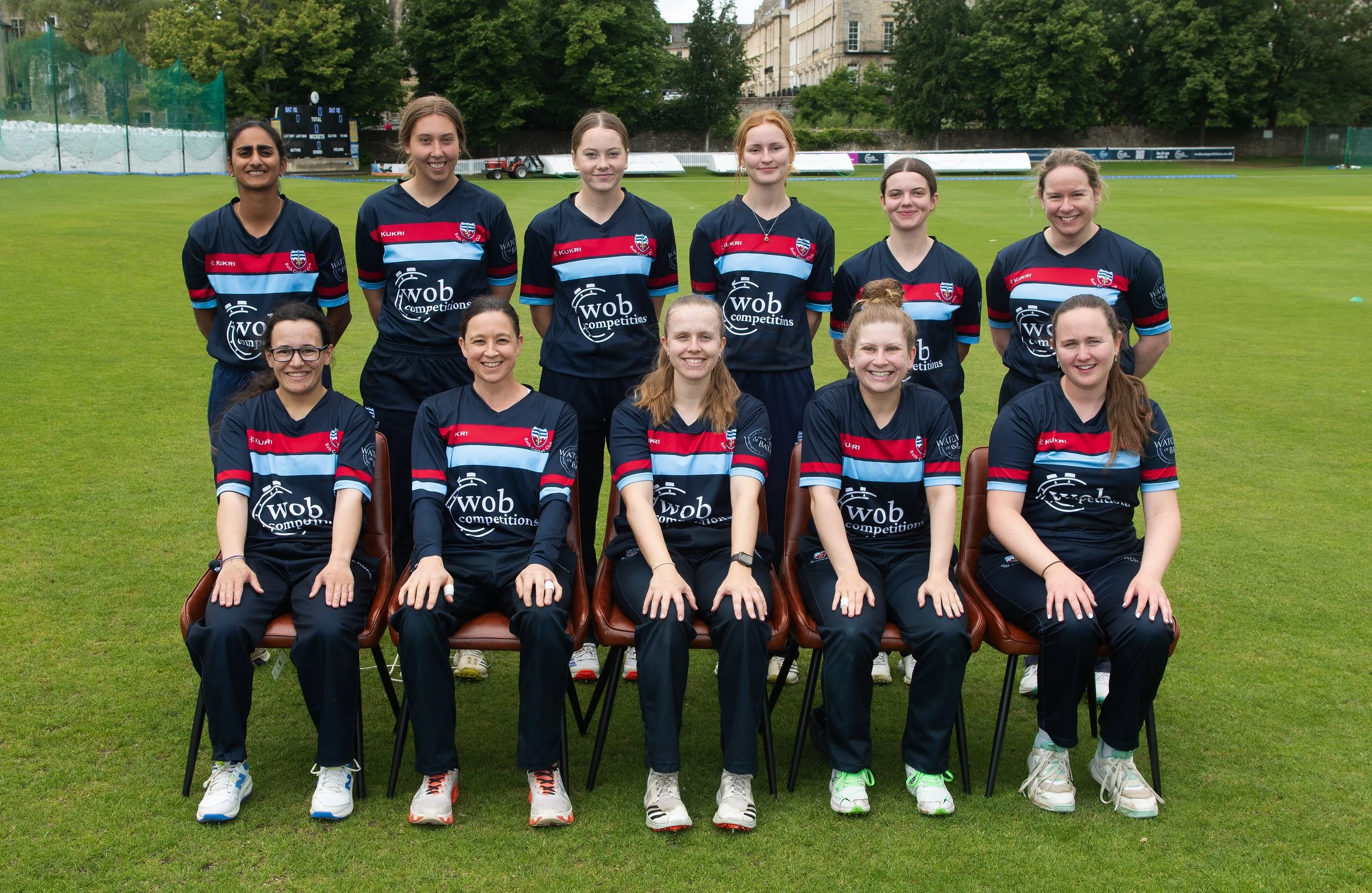 A women's sports team poses for a group photo on a grassy field, with some seated and others standing behind them. They are wearing matching navy sports jerseys with red, light blue, and white stripes and logos. Buildings and trees are visible in the background.