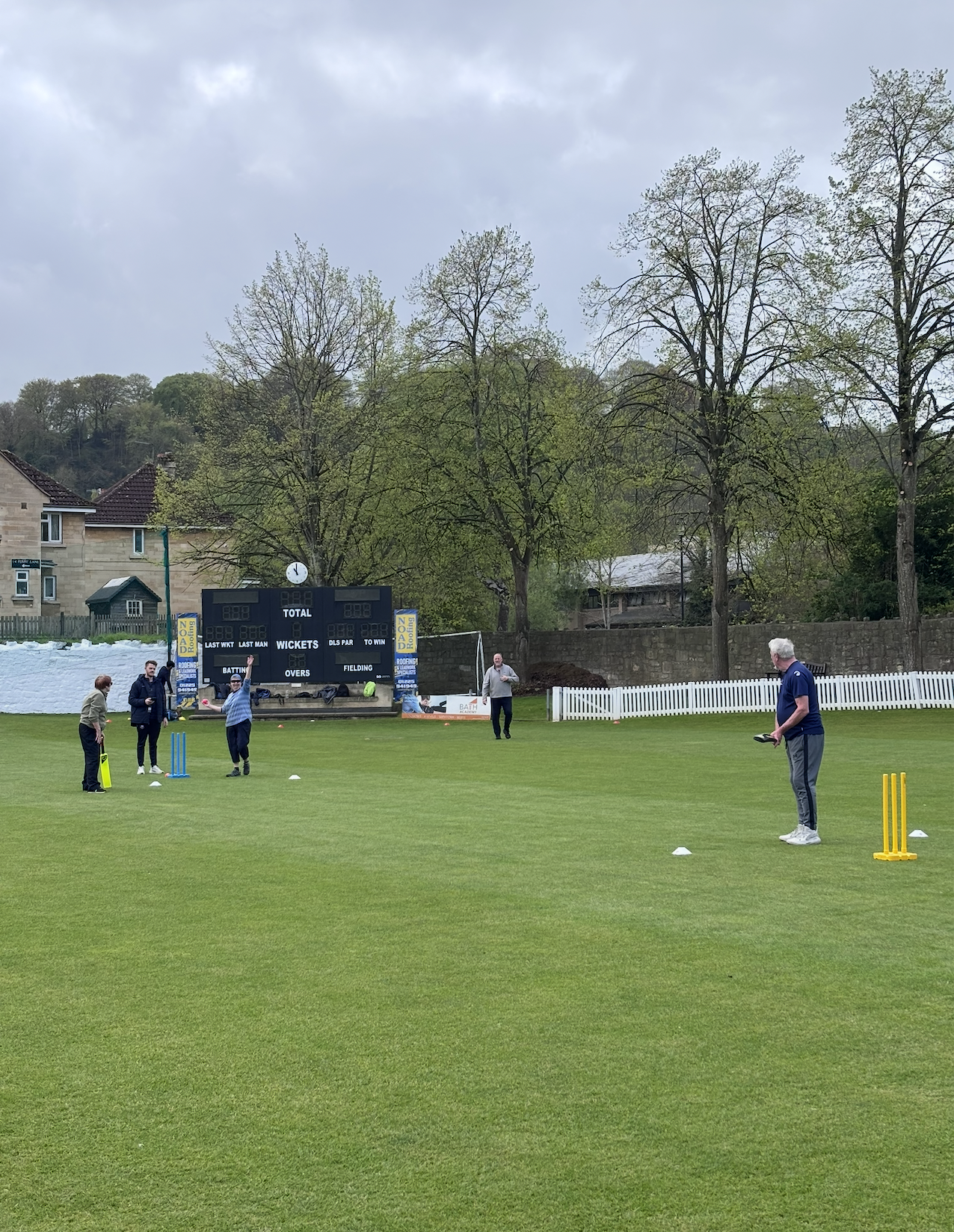 People practicing cricket on a grassy field with a scoreboard in the background and trees around.