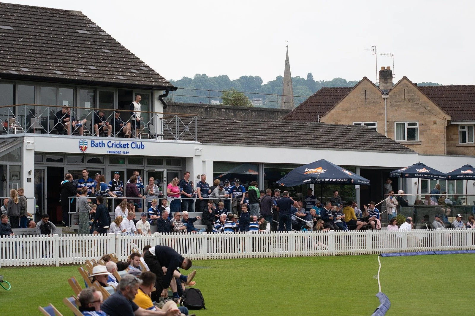 People watching a cricket game at Bath Cricket Club, with spectators sitting and standing near the field, and some on the upper balcony. The cricket field is enclosed by a white picket fence, with residential houses and a church steeple in the backgr