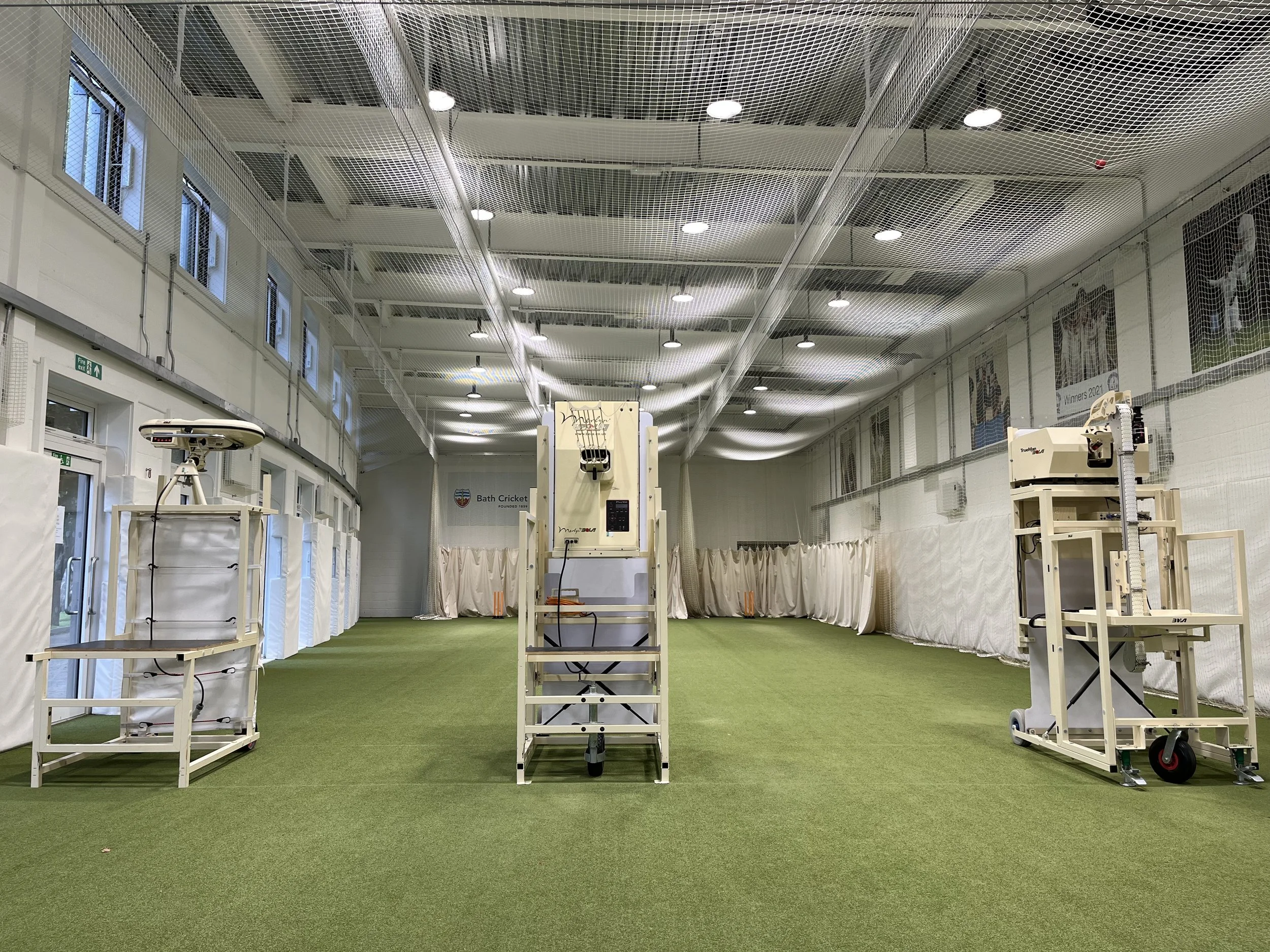 Indoor sports facility with green artificial turf, several robotic bowling pin setters, and a basketball hoop, enclosed by netting and large windows.
