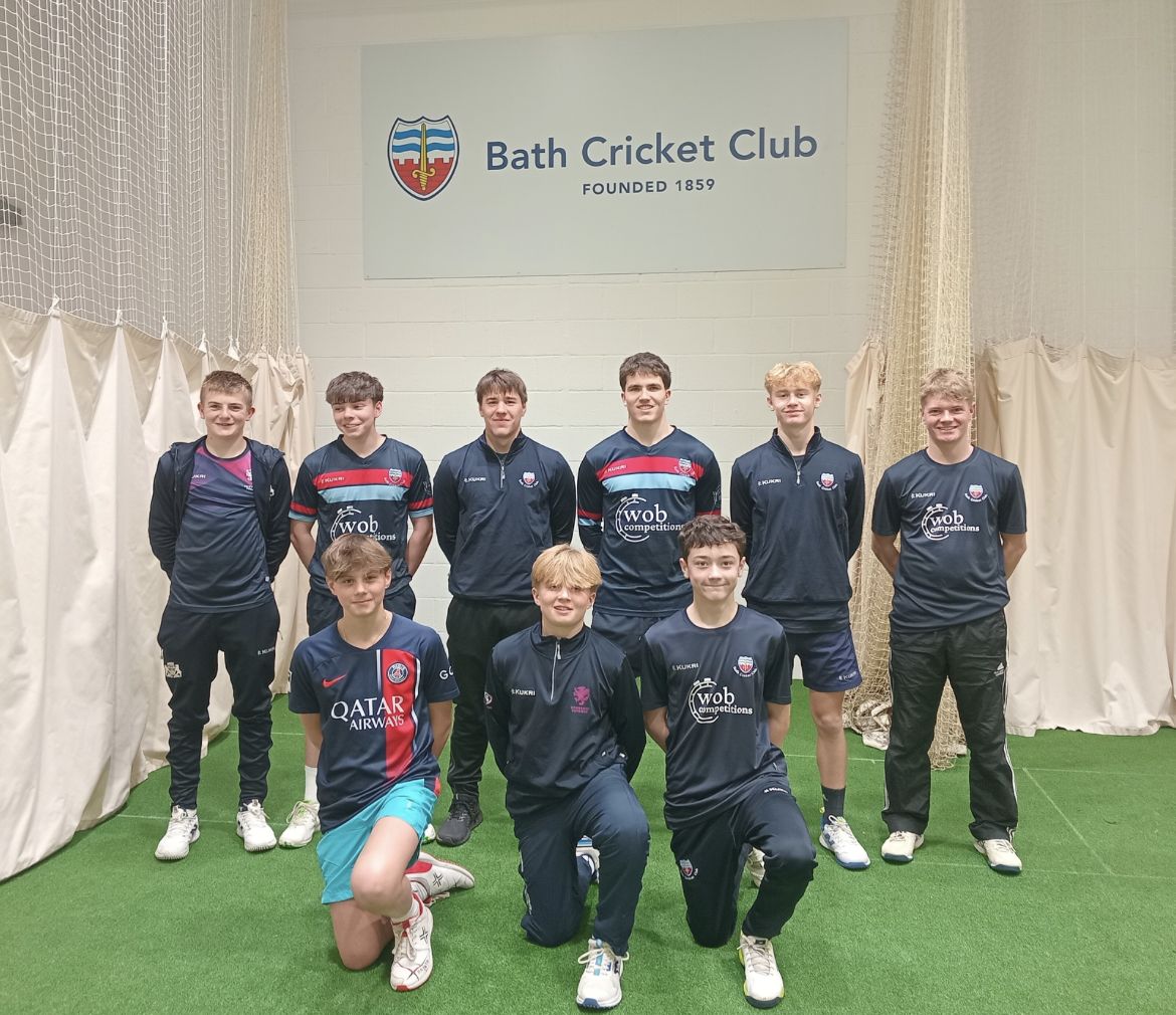 Group of young male cricket players in team uniforms posing inside Bath Cricket Club, founded 1859, with a sign on the wall.