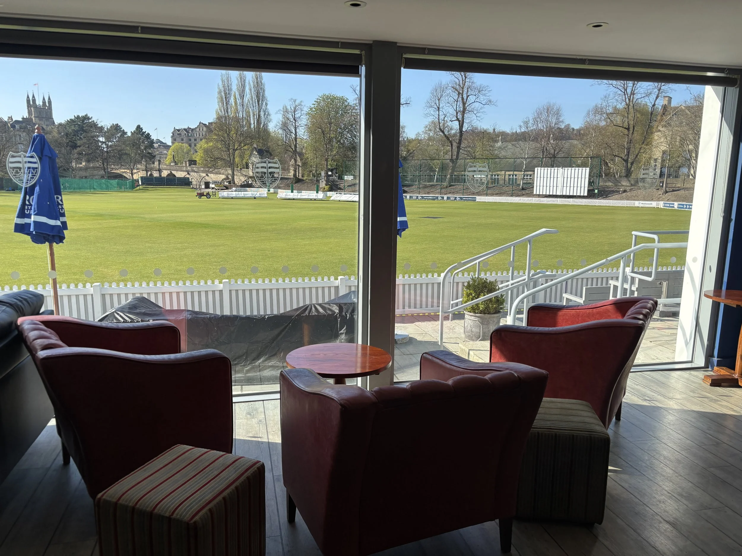 Indoor view of a sports field with green grass, surrounded by trees and buildings, seen through large glass windows with trays and chairs inside.