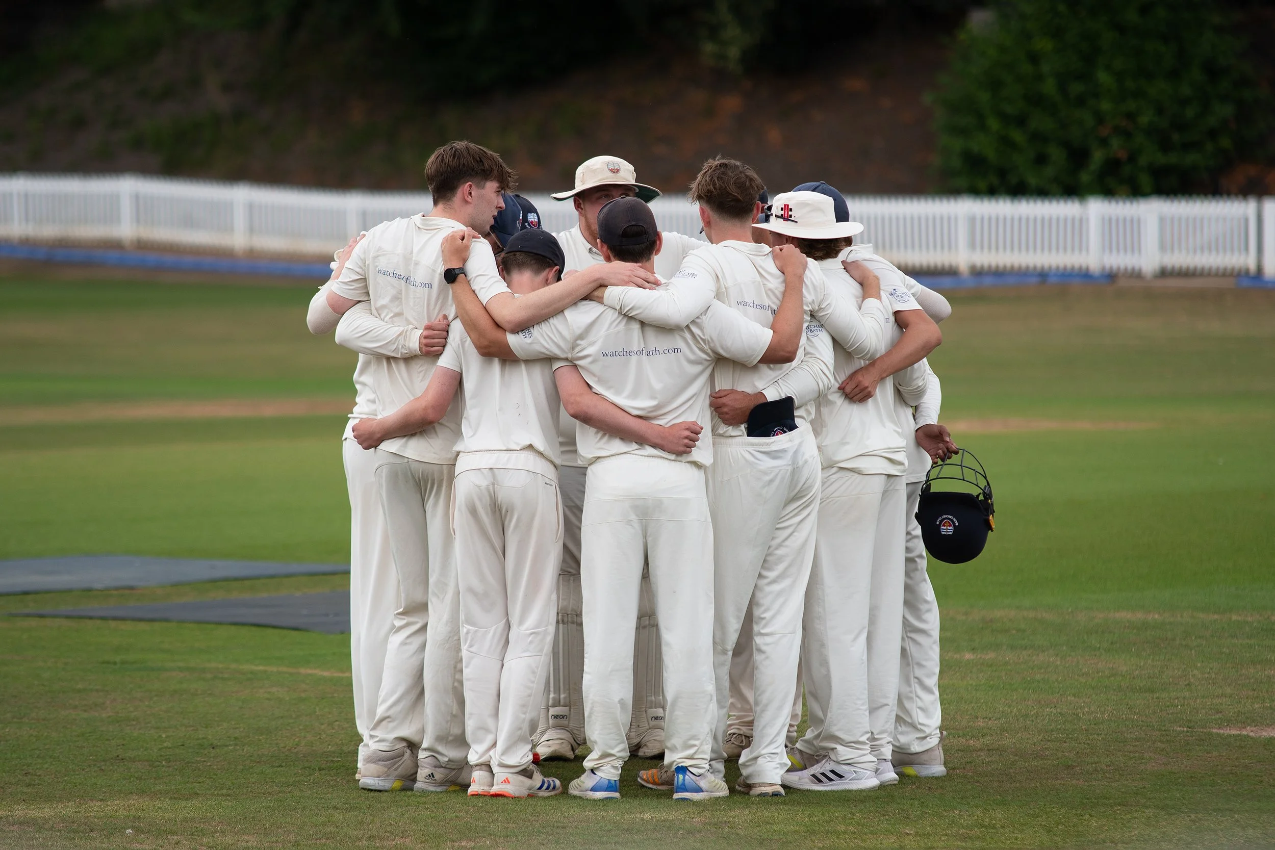 A group of cricket players in white uniforms huddle on a cricket field, embracing each other in a team cheer.