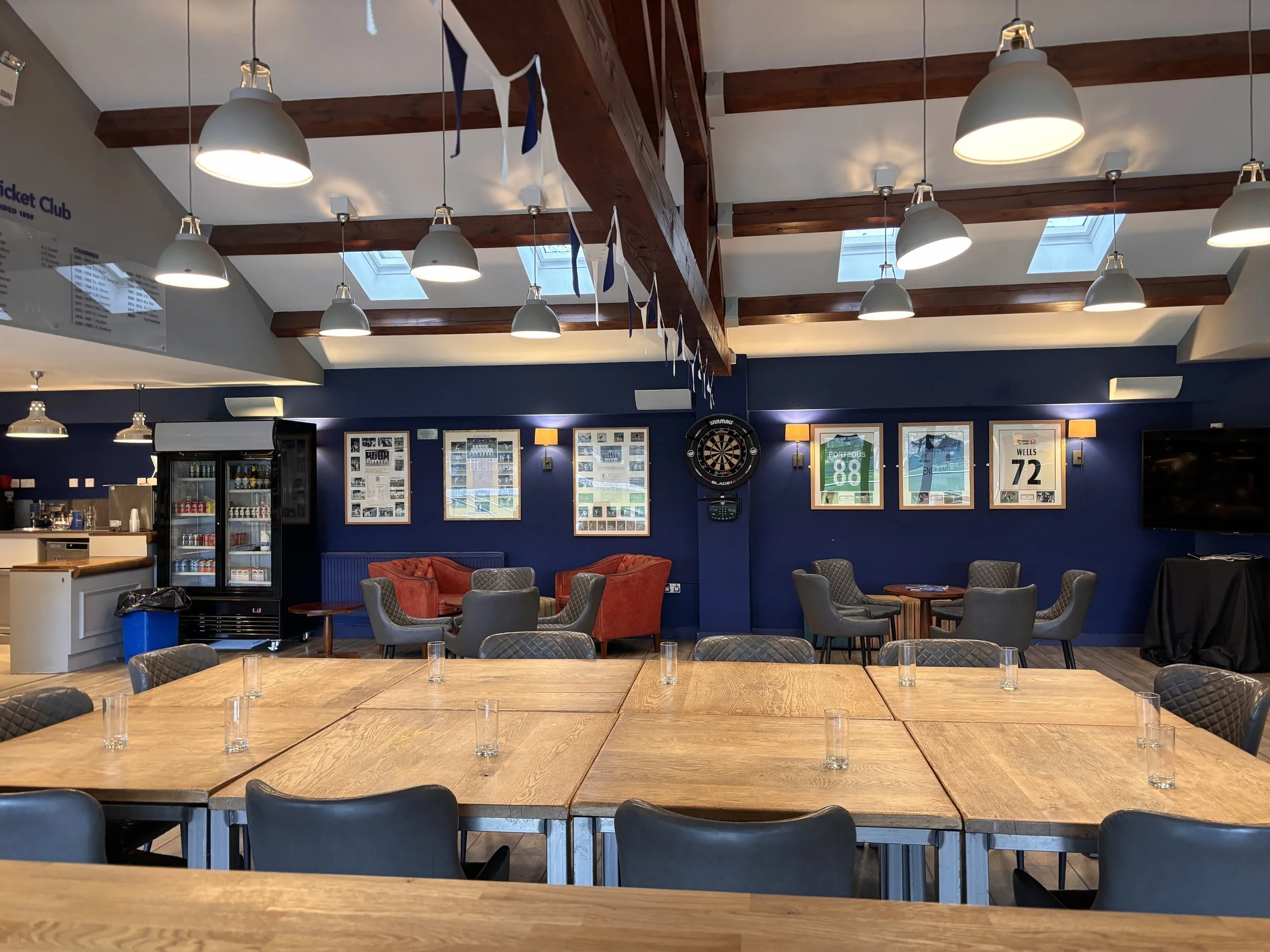Interior of a modern recreational room or club with wooden ceiling beams, multiple hanging lights, framed sports posters on the navy blue wall, a dartboard, a small fridge, and chairs around large wooden tables, illuminated by skylights.