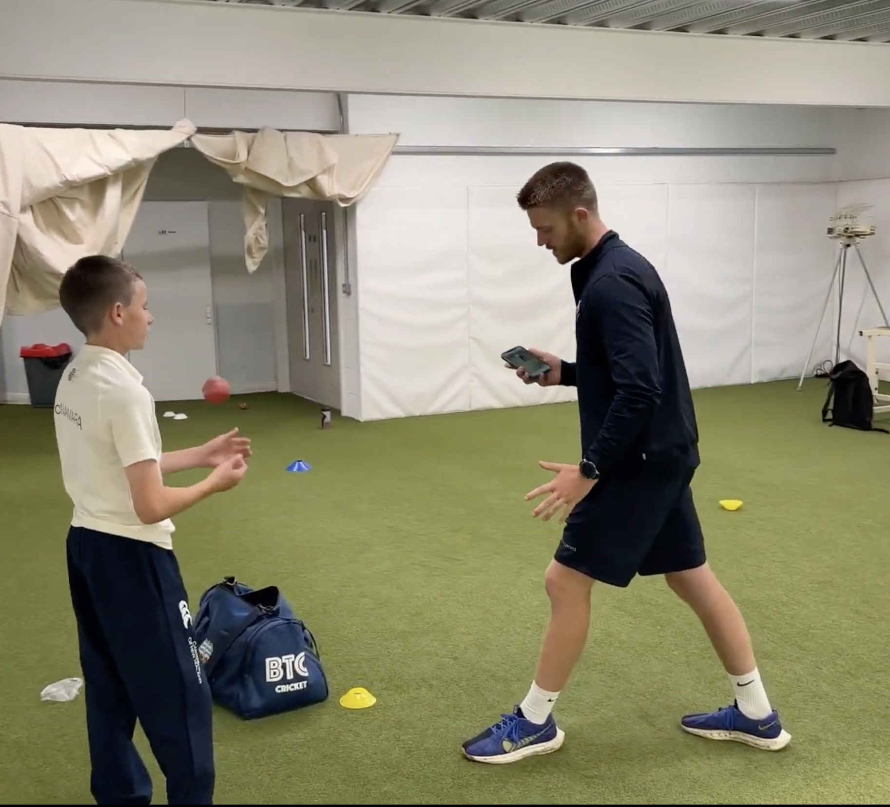 A cricket coach demonstrating a technique to a young player in an indoor practice facility. The coach is wearing a black jacket and shorts, holding a phone, while the boy in a white sports shirt and navy pants is preparing to bowl, surrounded by sports equipment and training markers on the floor.