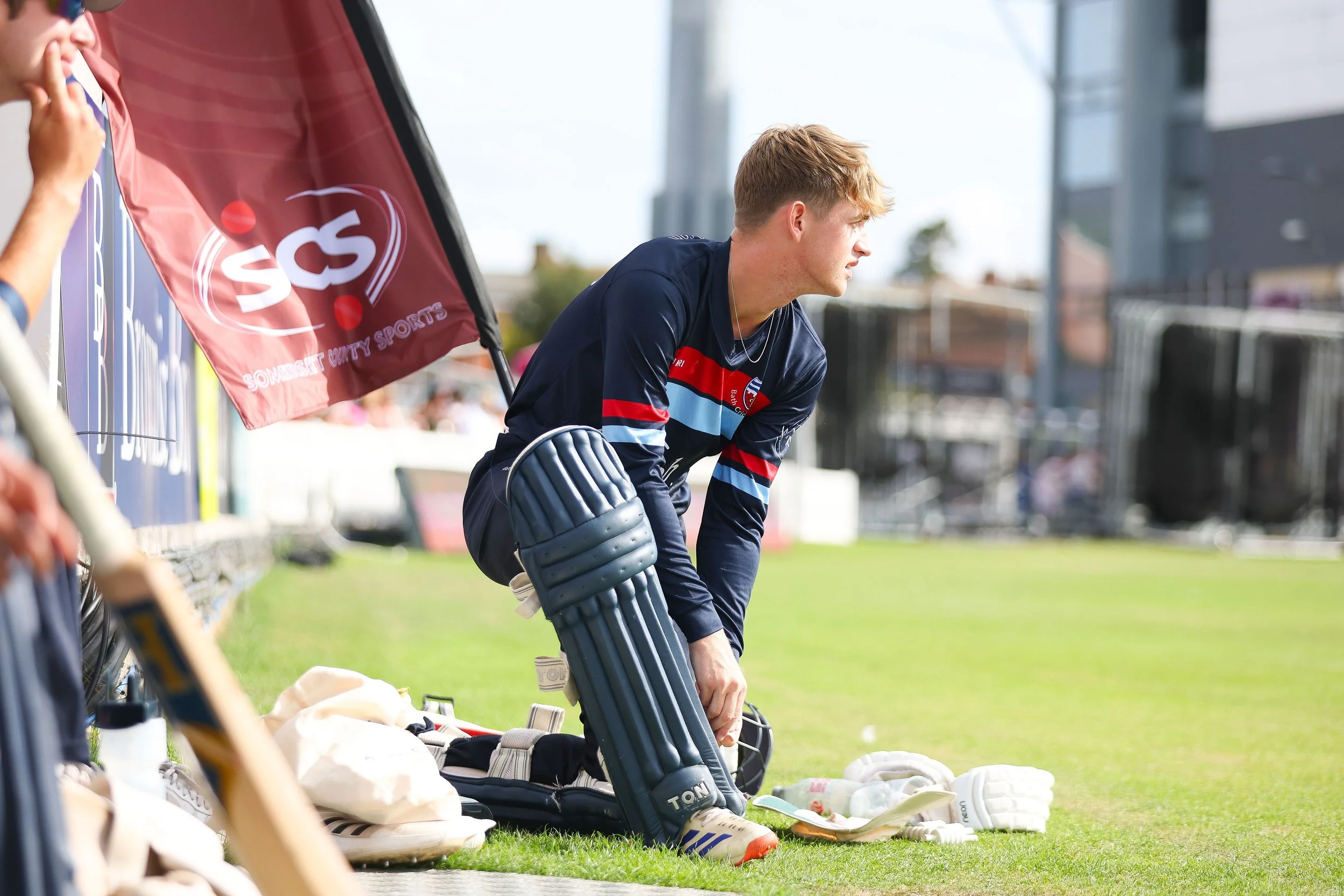 A young male soccer player in a navy and red jersey kneeling on the sidelines, resting with his hand on his knee, surrounded by soccer equipment including gloves and shoes.