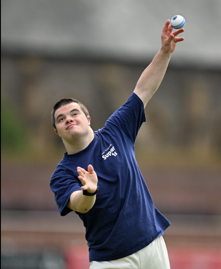 A man in a navy blue t-shirt with white text throws a ball in the air outdoors.