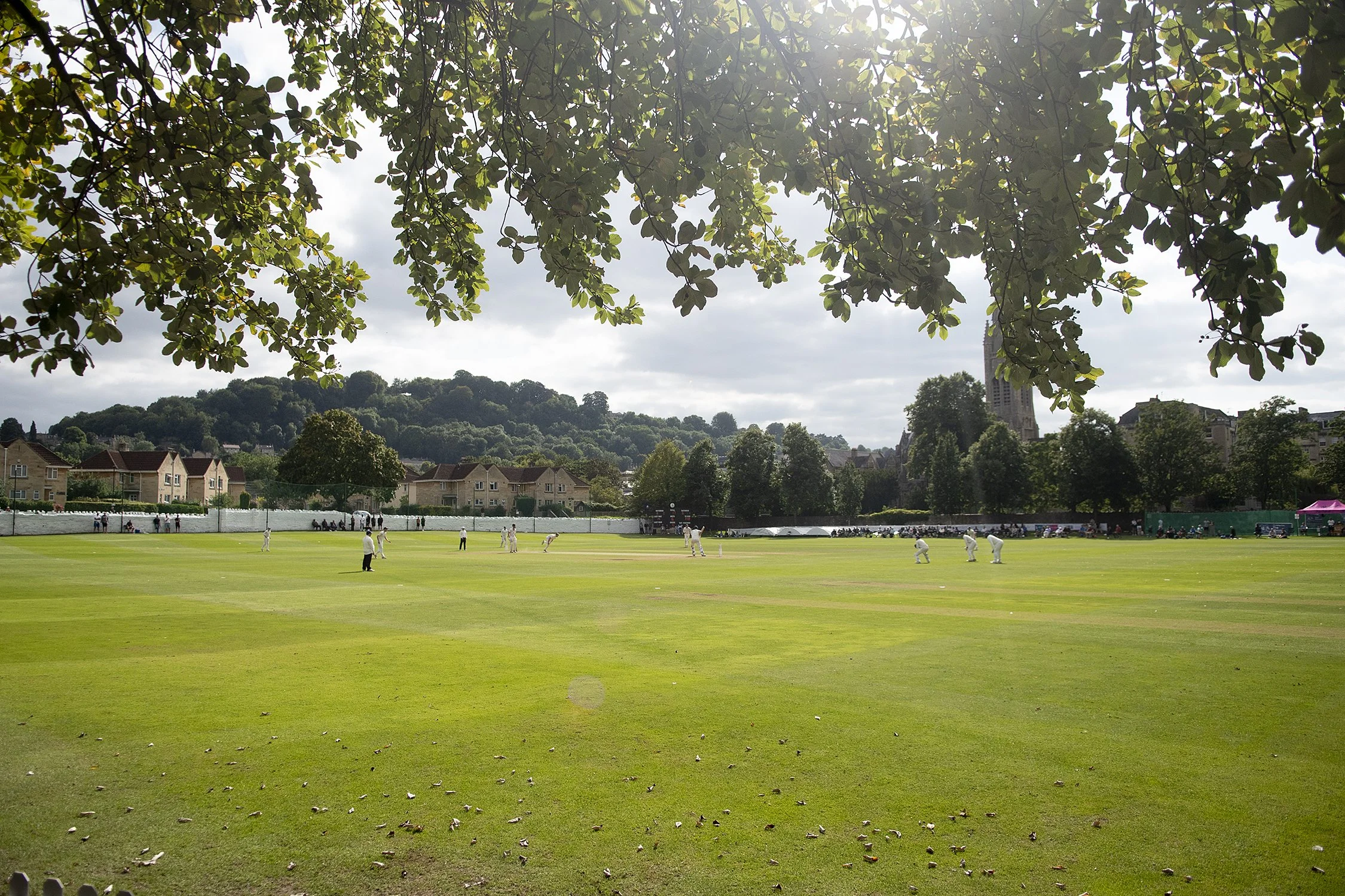 Cricket players on a green field, with houses and a church tower in the background, under a partly cloudy sky with sunlight filtering through tree branches overhead.