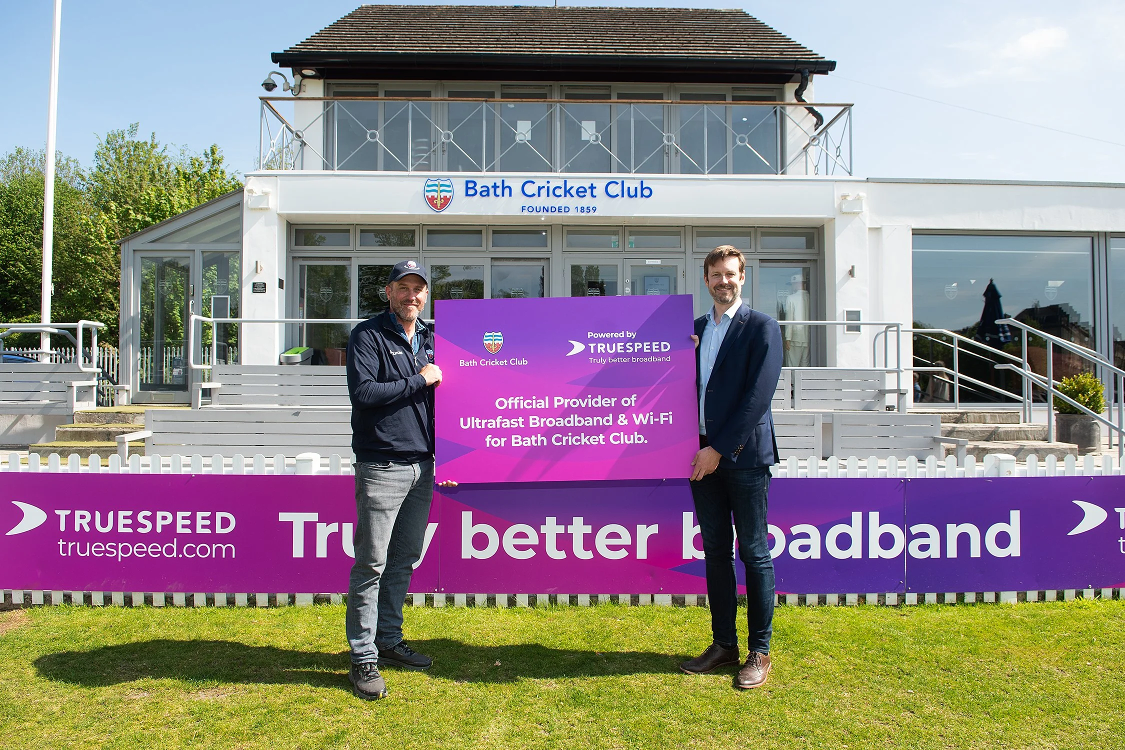 Two men are standing in front of a purple sign at the Bath Cricket Club, holding a sign that reads 'Powered by TRUEPSPEED, Official Provider of Ultrafast Broadband & Wi-Fi for Bath Cricket Club.' The building behind them has a sign that says Bath Cricket Club, founded in 1859.