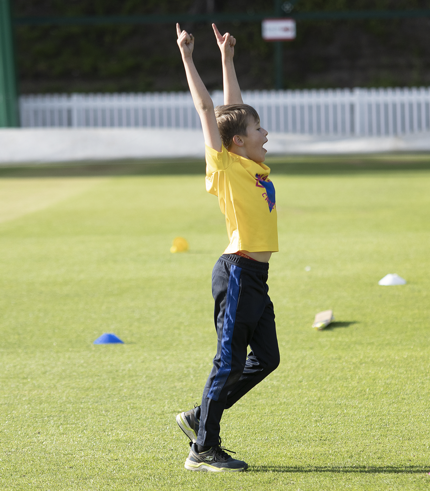 Young boy in a yellow shirt and black pants on a grass field with cones, cheering with arms raised.