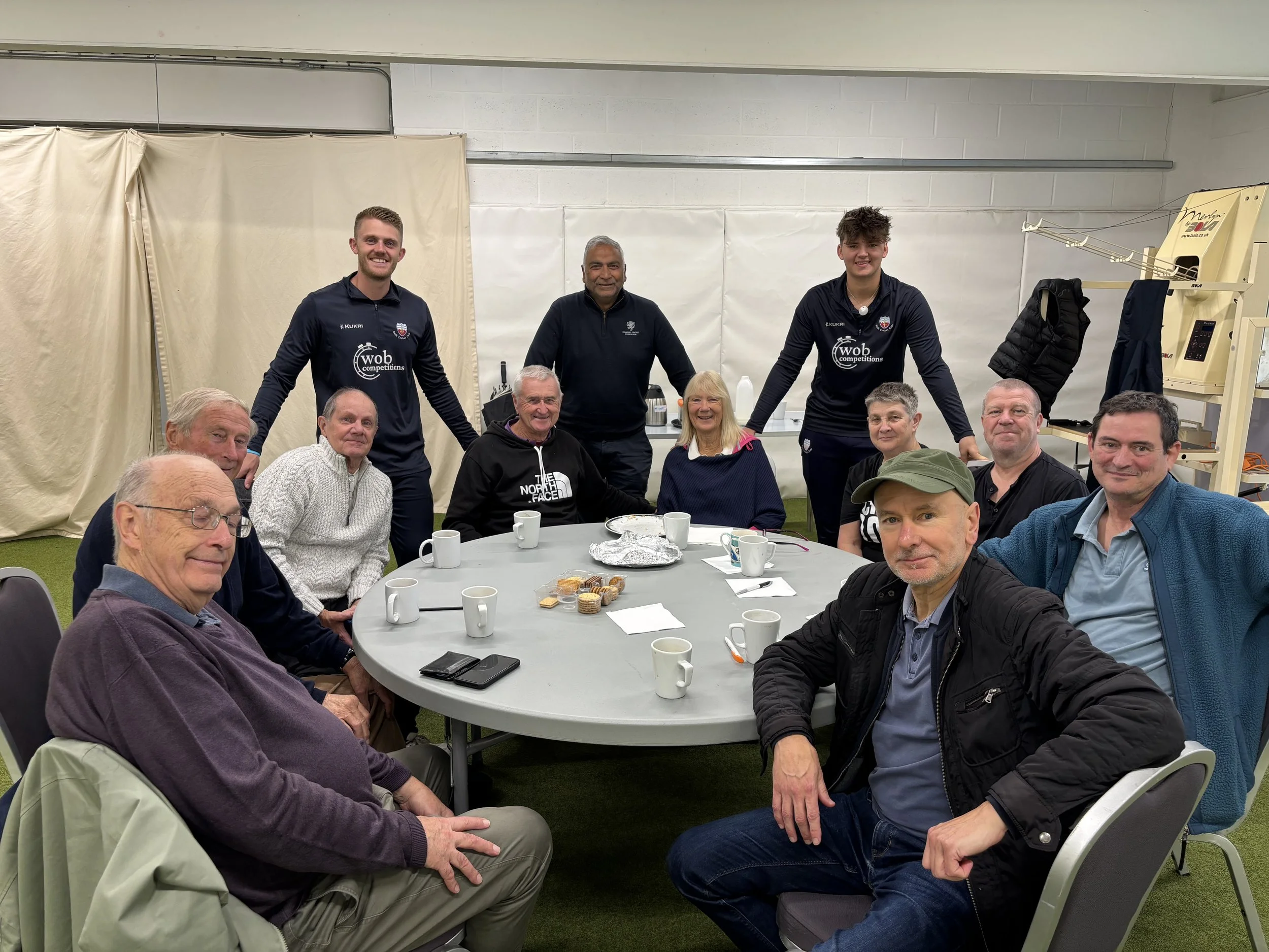 A group of older and younger men and women sitting and standing around a table, smiling, with snacks and drinks, in an indoor setting with white walls and beige curtains.