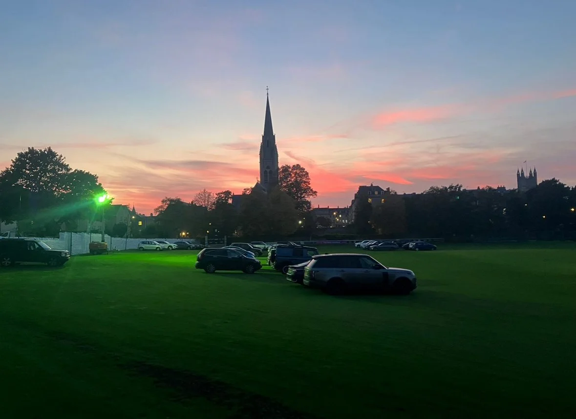 A parking lot with several cars on green grass, a church steeple against a colorful sunset sky, and some trees.