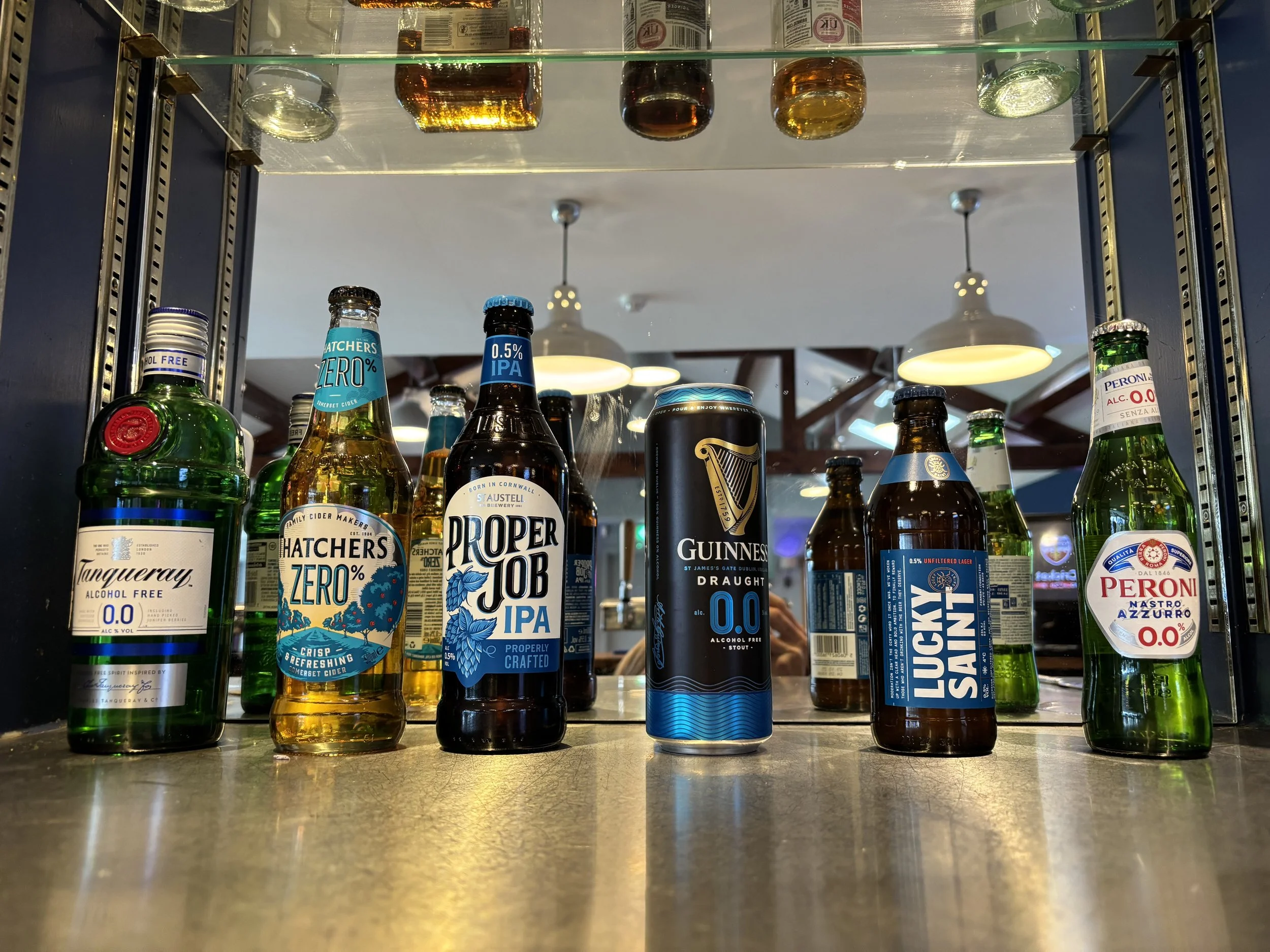 A refrigerator glass door shows various non-alcoholic and alcoholic beverages, including beer bottles, cans, and cider bottles, with a mirror-like reflection and a ceiling with hanging lights in the background.