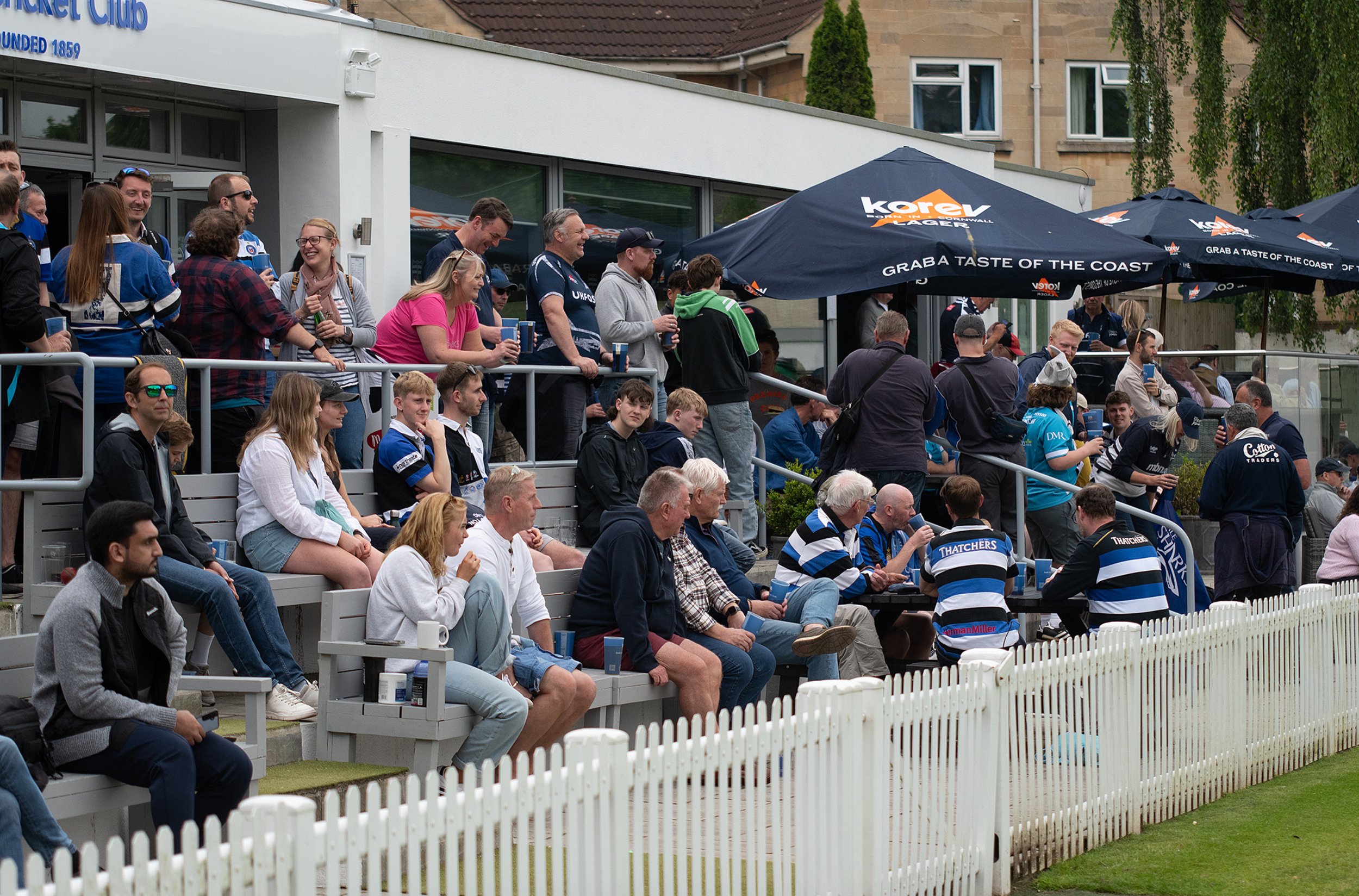 Crowd of people sitting and standing in outdoor stadium seating, with some wearing rugby team jerseys and others holding drinks. There are umbrellas with advertising and a white picket fence in the foreground.