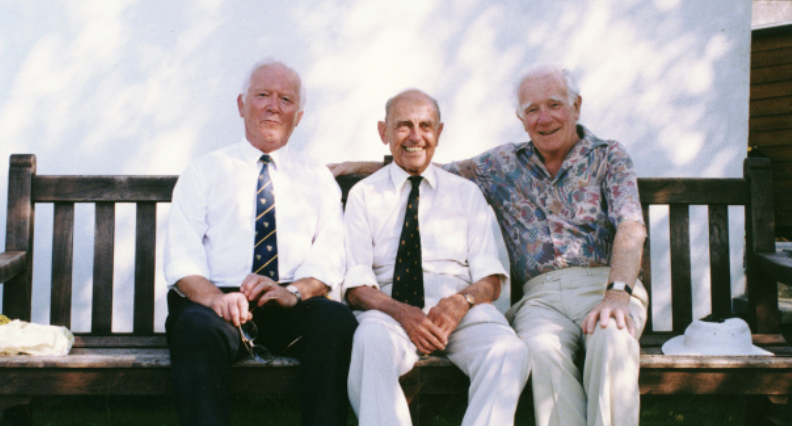 Three elderly men sitting on a wooden bench outdoors, smiling, with a partly cloudy sky in the background.