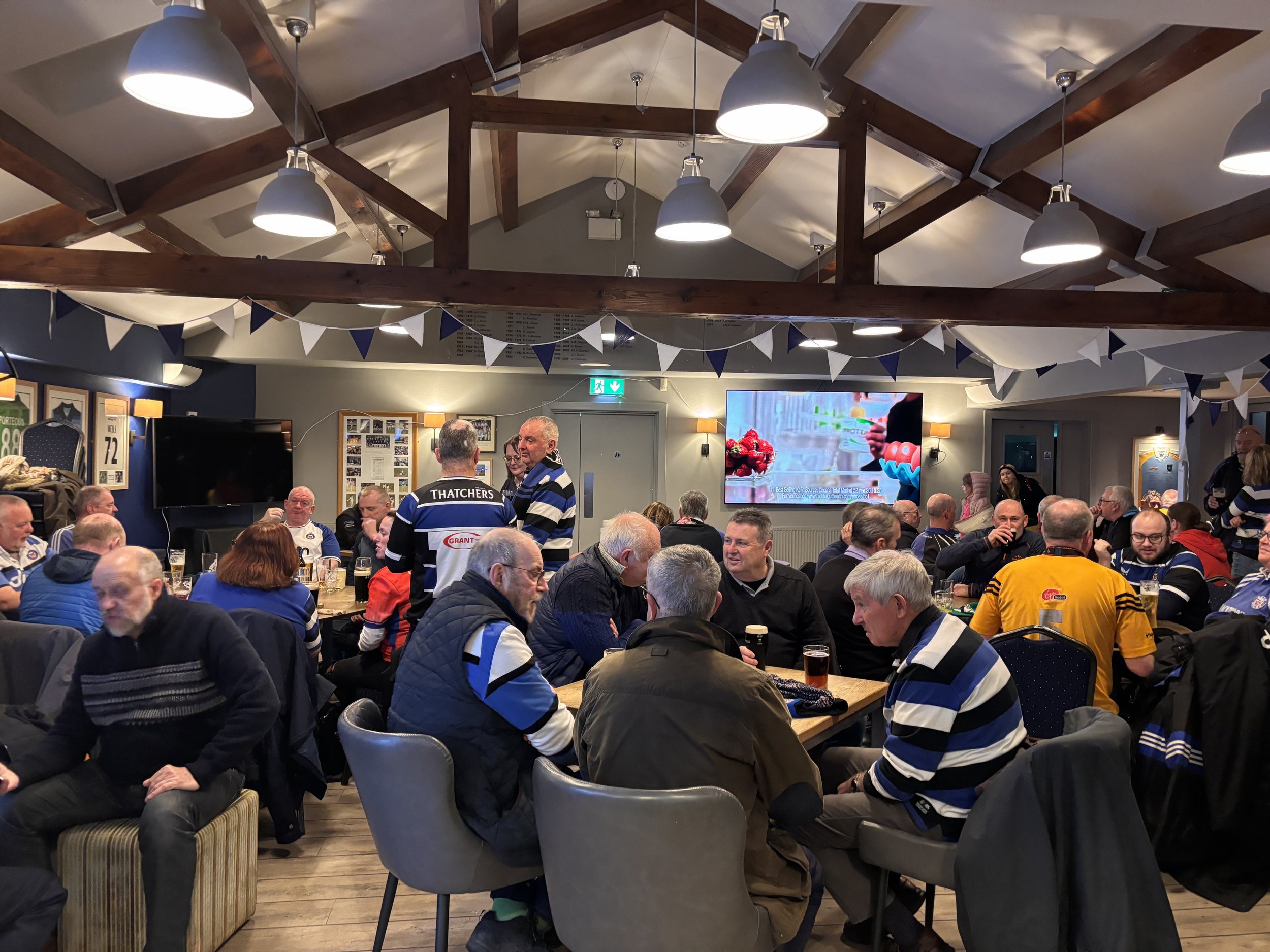 A large group of people gathered in a cozy, well-lit room with wooden beams and dark blue walls. Many attendees are wearing sports jerseys, some are talking, and others are sitting at tables. The room is decorated with bunting and has a large screen 