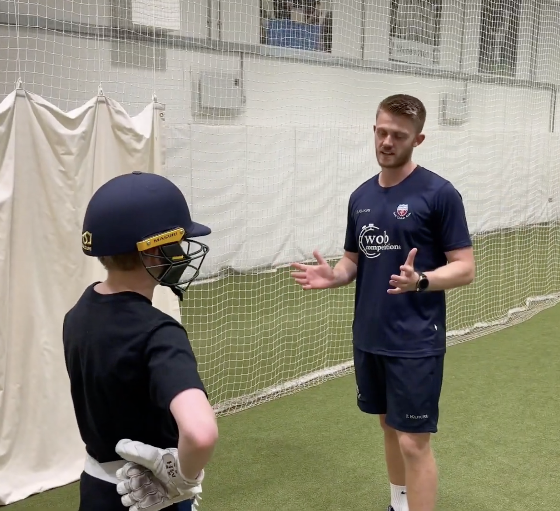 A young man wearing sports attire, talking to a child in sports gear, inside an indoor sports facility with a netted background.