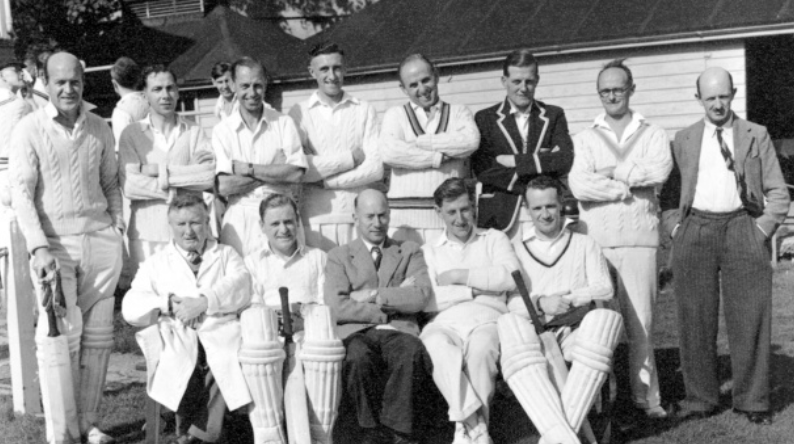 Black and white photo of a group of eleven men, most wearing cricket uniforms, posing outdoors in front of a building. Four are seated, and the others are standing behind them.