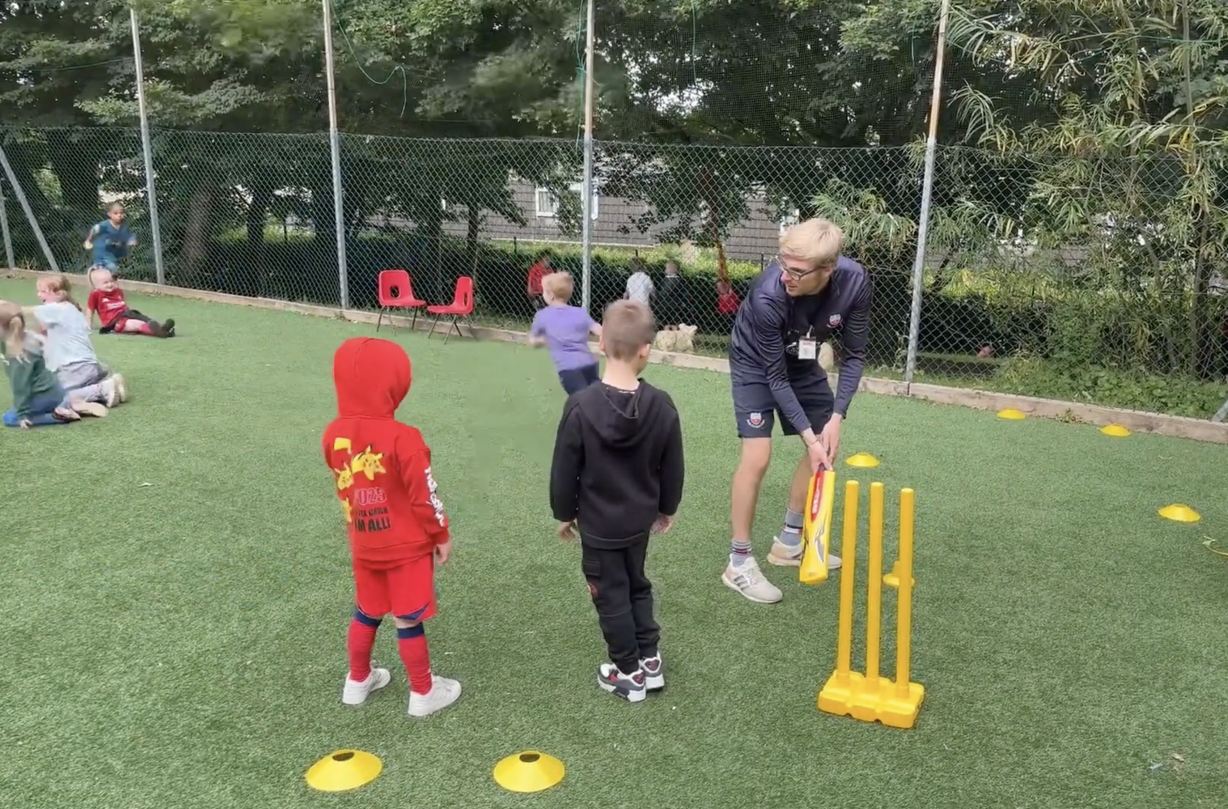 Children playing outdoors on a grassy field with a young man, likely a coach, guiding them. There are kids sitting and running, some dressed in colorful sportswear. Yellow cones are used for activities, and a chain-link fence surrounds the area with trees in the background.