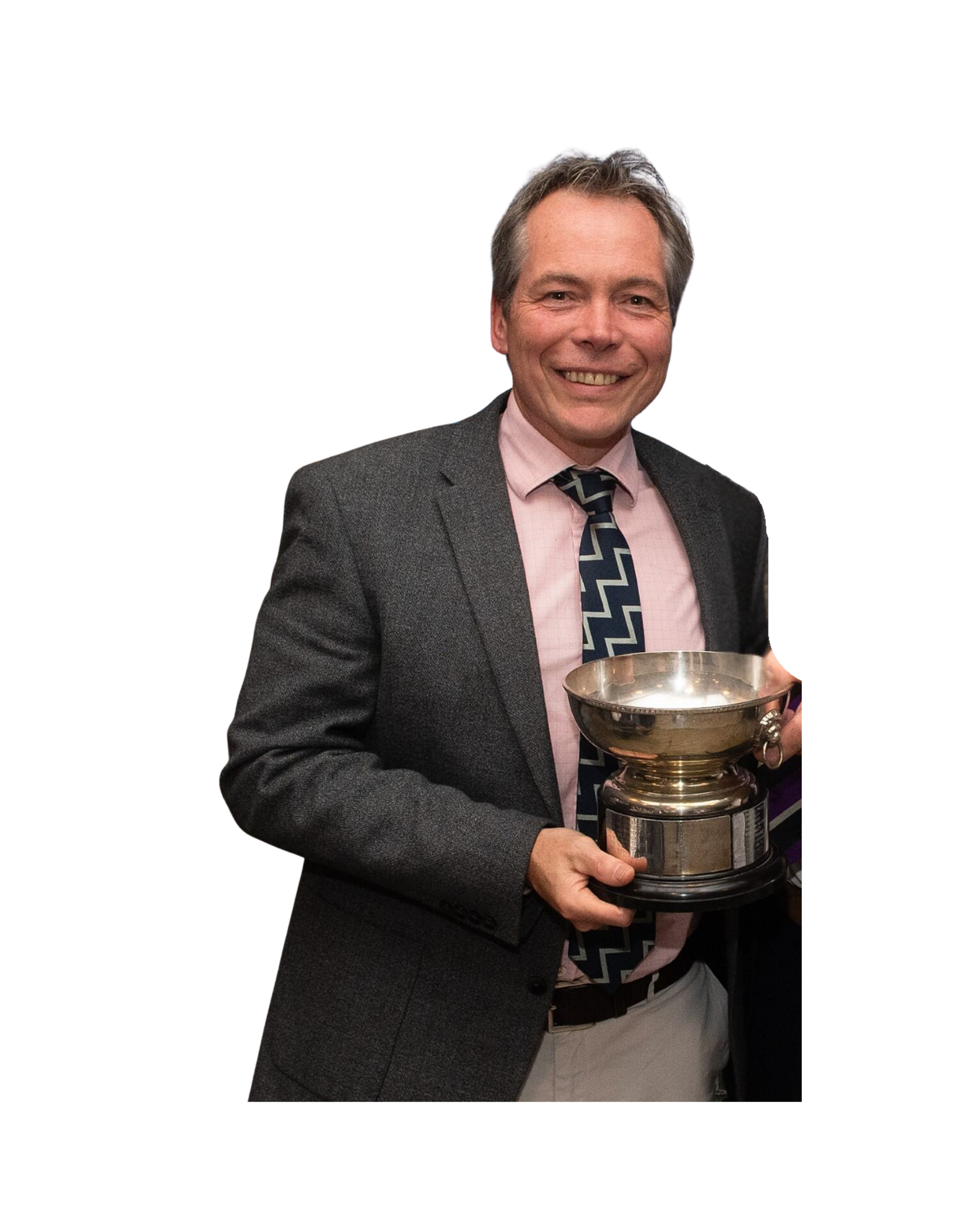 A man with gray hair and a big smile, wearing a suit jacket, pink shirt, and a patterned tie, holding a large silver trophy cup.