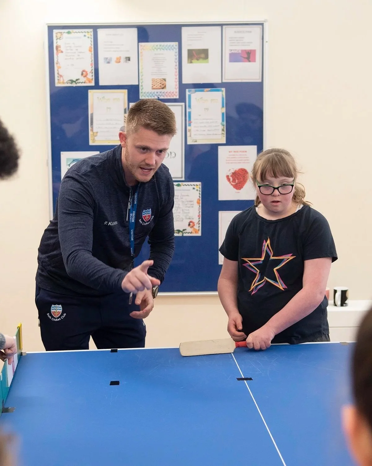 A man and a girl playing table tennis indoors, with a blue background bulletin board behind them.