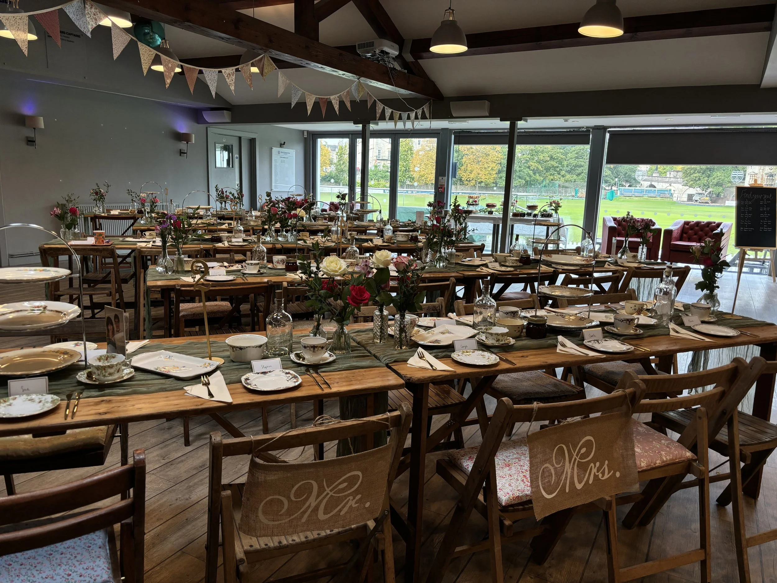 Interior of a decorated event space with tables set for a meal, floral centerpieces, bunting decorations hanging from the ceiling, and large windows with a view of a park outside.