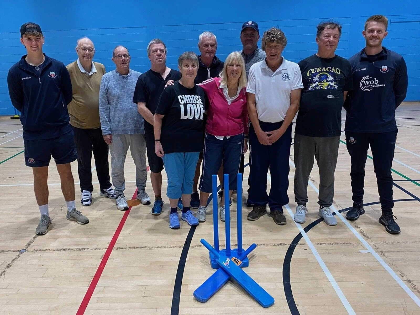 Group of eleven people standing in a sports gymnasium with cricket stumps in the foreground.