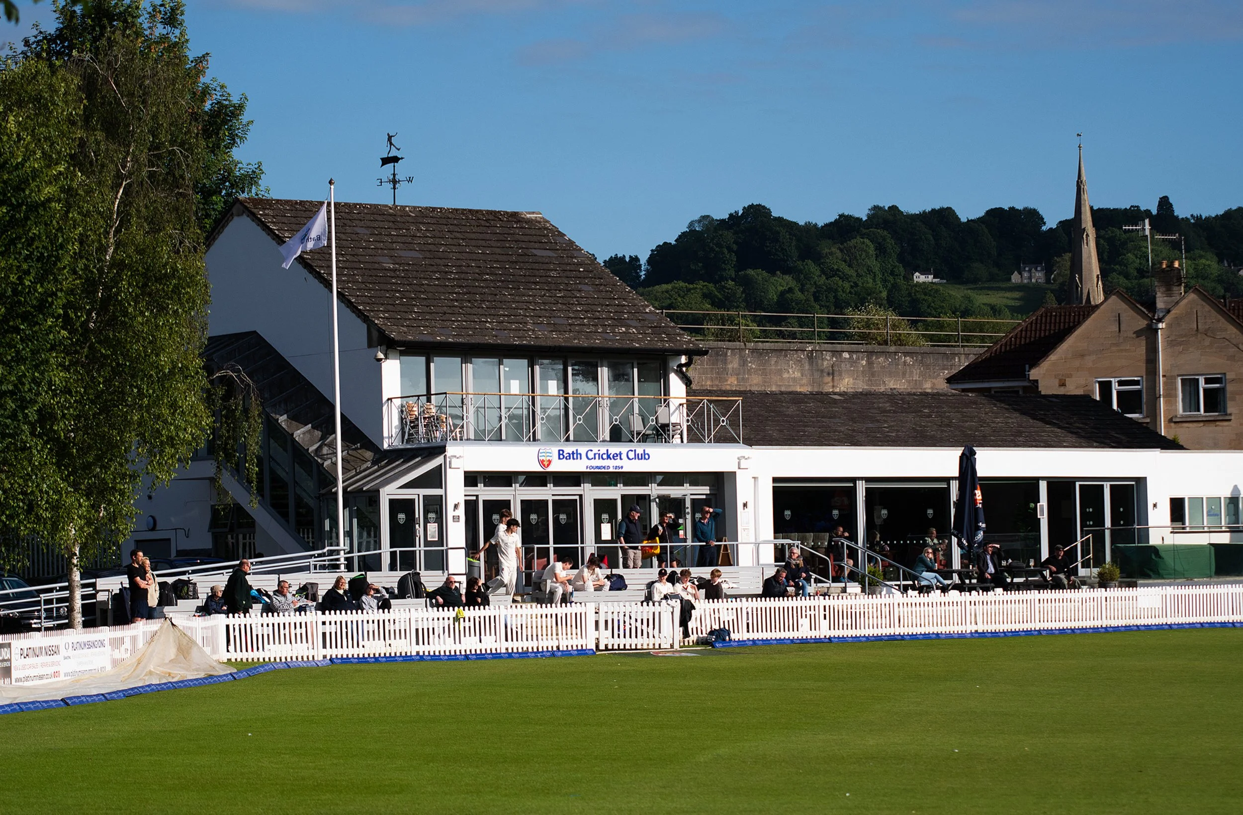 A cricket stadium with spectators seated along the field, a white building labeled Bath Cricket Club, and a grassy playing field in the foreground. In the background, there are trees, a hill with houses, and a church steeple.