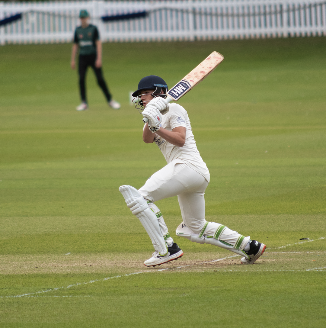Cricket player in white uniform running on the field with bat in hand, wearing helmet and pads, while an outfielder is in the background.