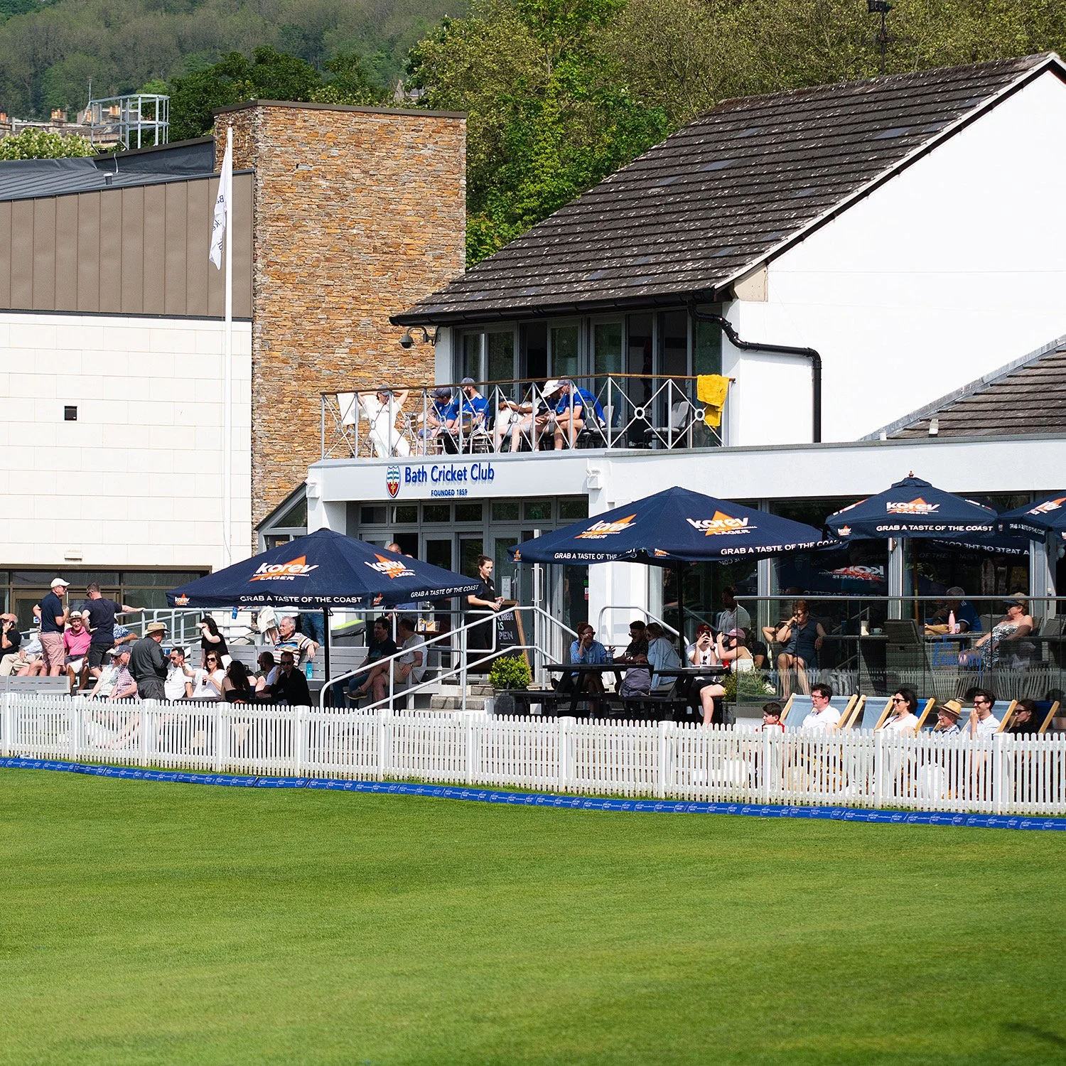 People watching a cricket match at Bath Cricket Club from a pavilion and outdoor seating area, with umbrellas, a white picket fence, and a grassy field in the foreground.