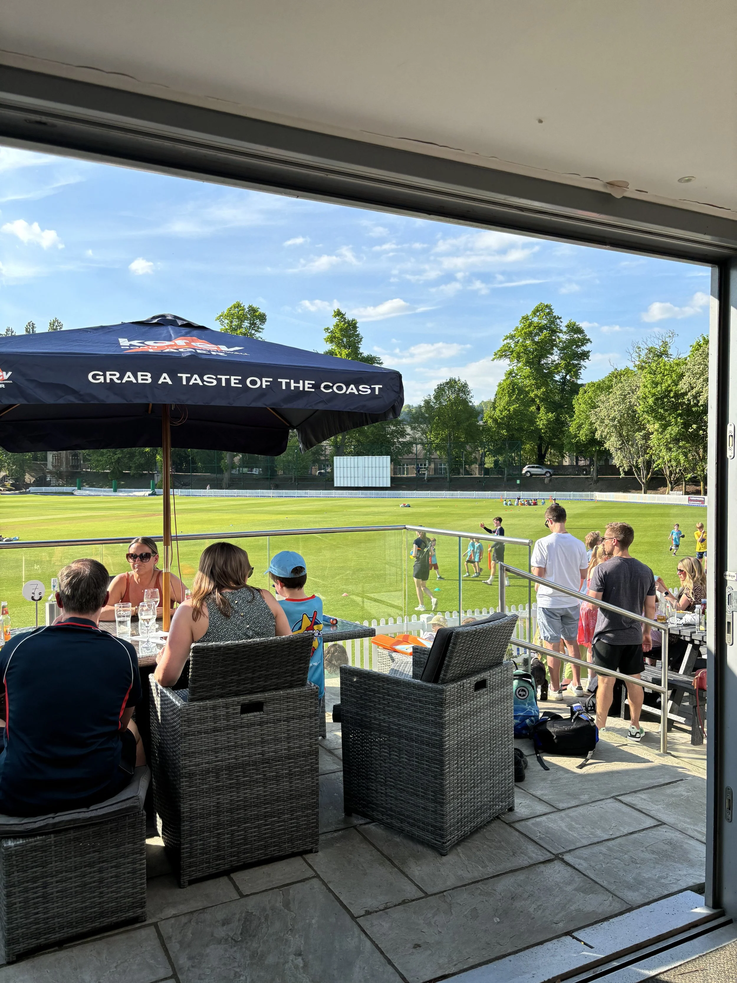 People sitting at a table under an umbrella on a patio overlooking a sports field, with players on the field and trees in the background.