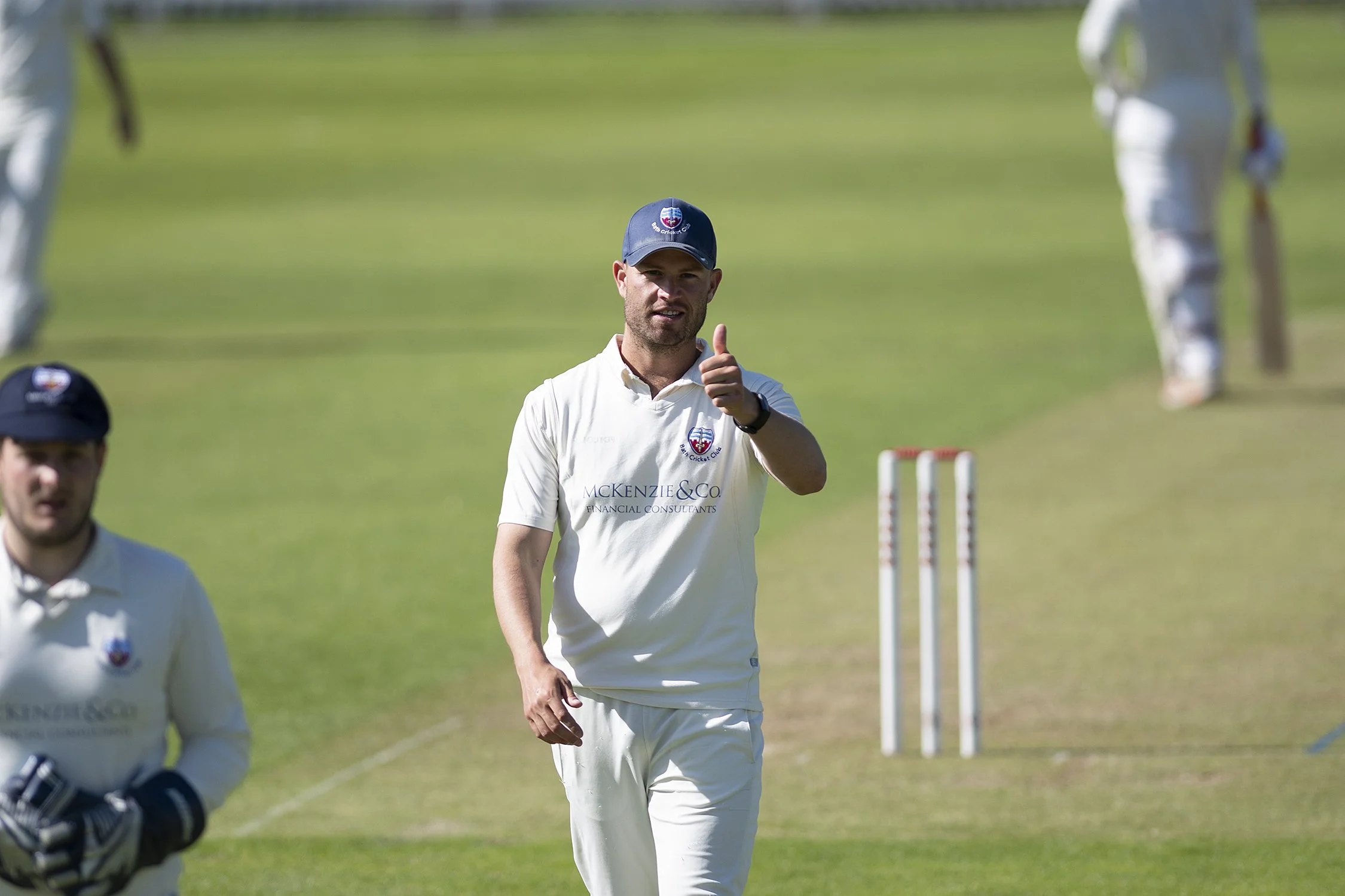 A man wearing a white cricket uniform and a blue cap giving a thumbs-up on a cricket field, with cricket stumps and other players in the background.