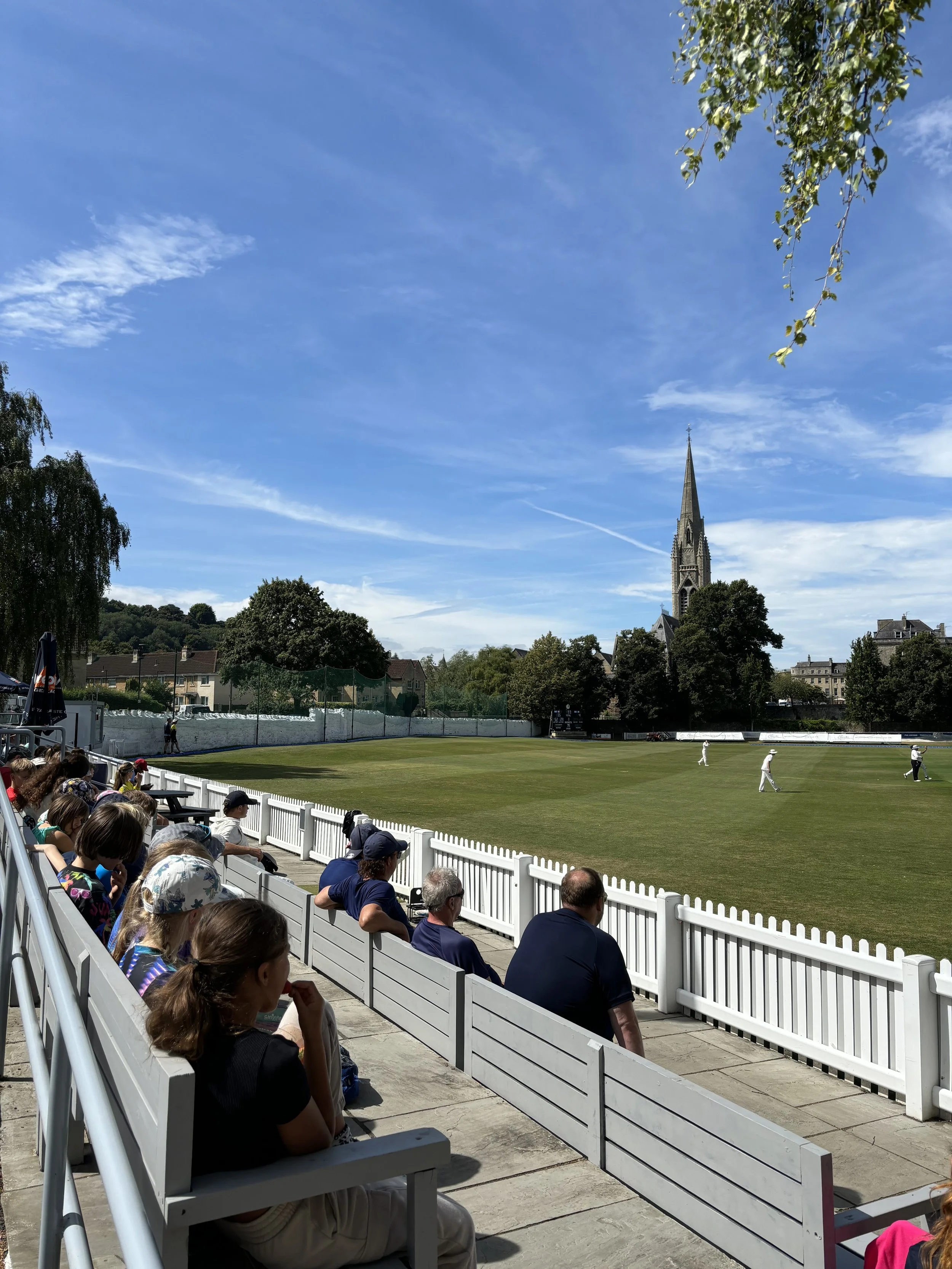 People sitting on white benches watching a cricket game on the field, with trees, some buildings, and a church with a tall spire in the background under a blue sky with some clouds.
