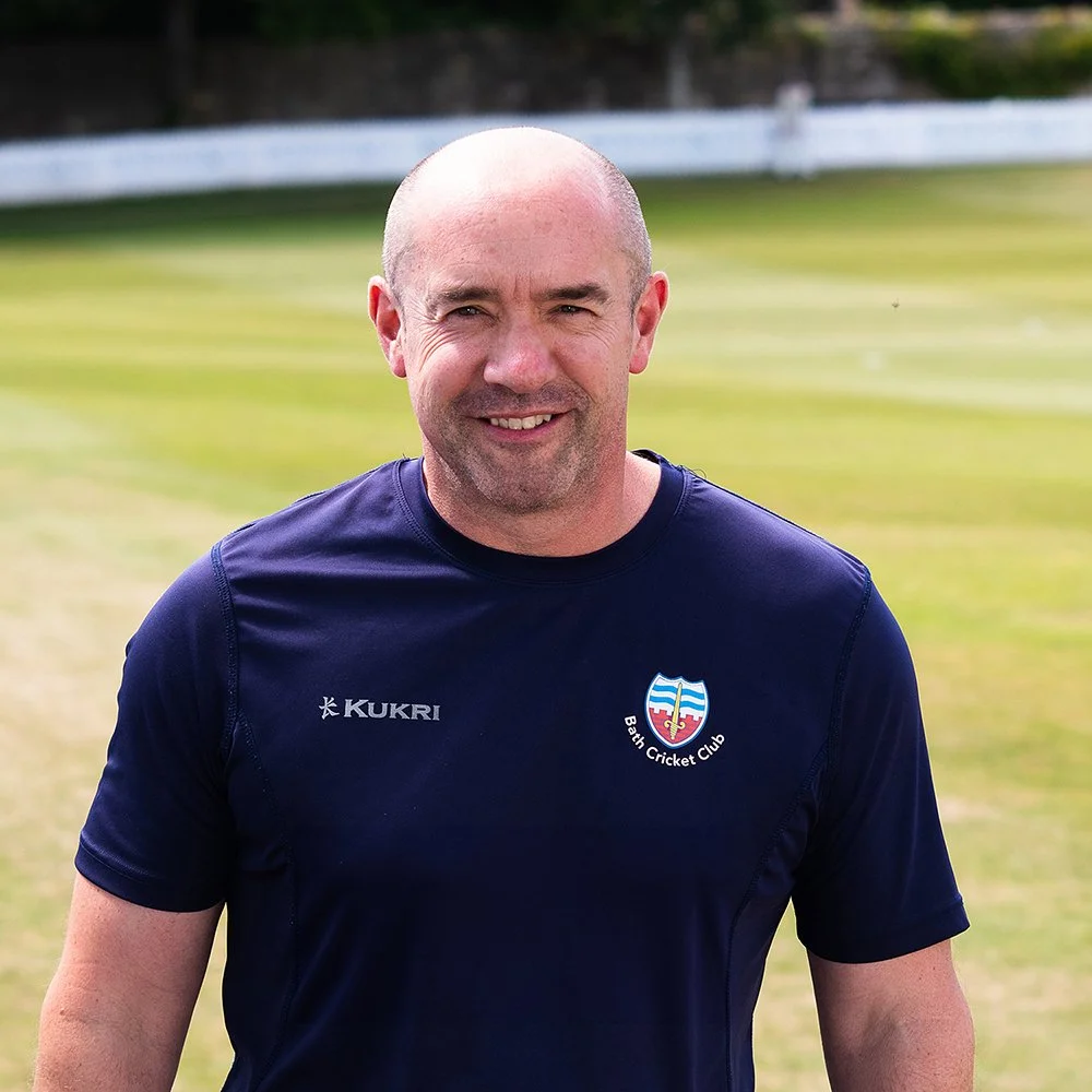 A smiling man standing outdoors on a cricket field, wearing a navy blue Bath Cricket Club shirt.