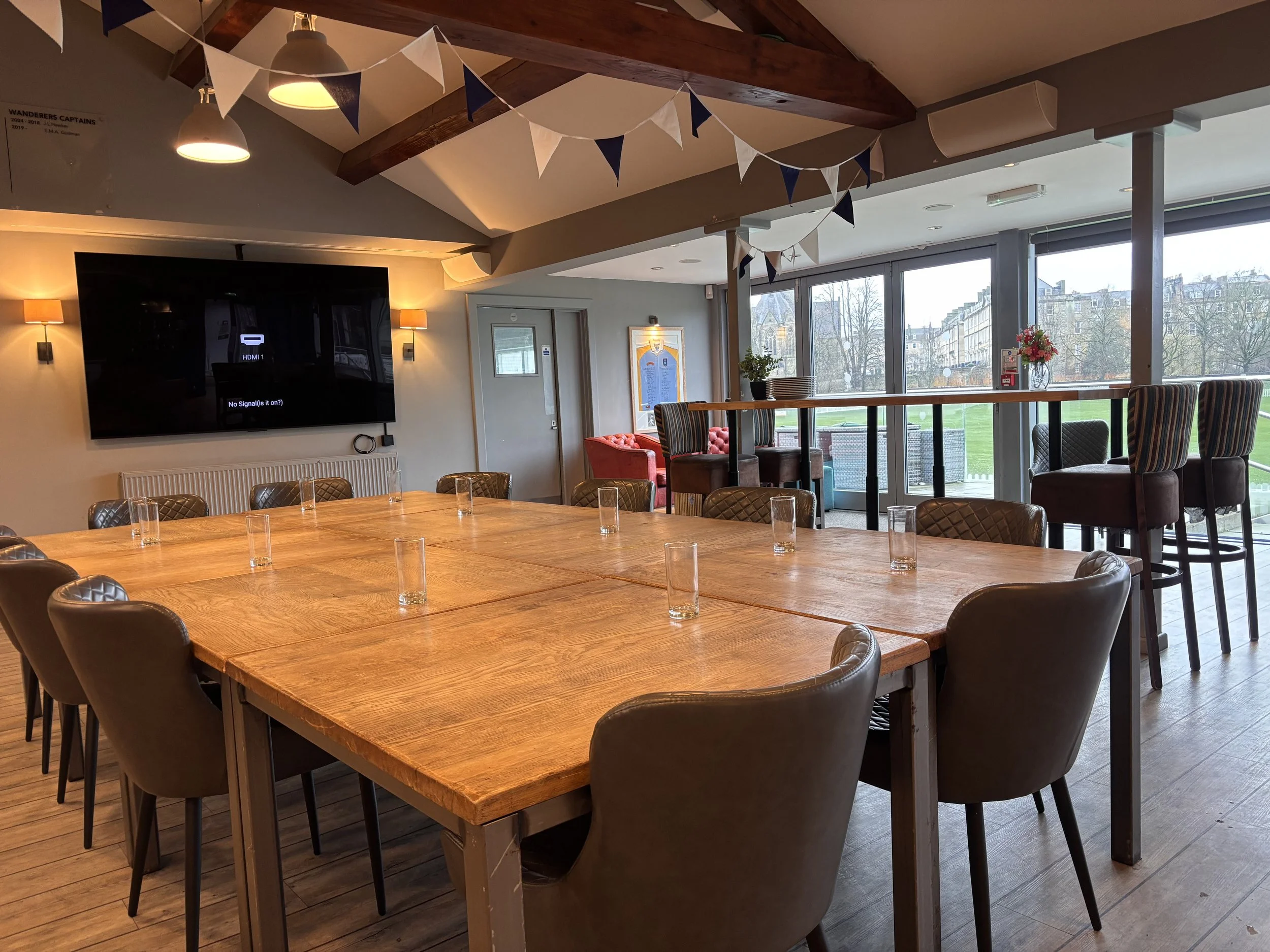 A spacious dining or meeting area with a large wooden table surrounded by brown leather chairs. The room has large windows letting in natural light and offering a view of a green outdoor space. Decor includes a string of white and blue pennant banner