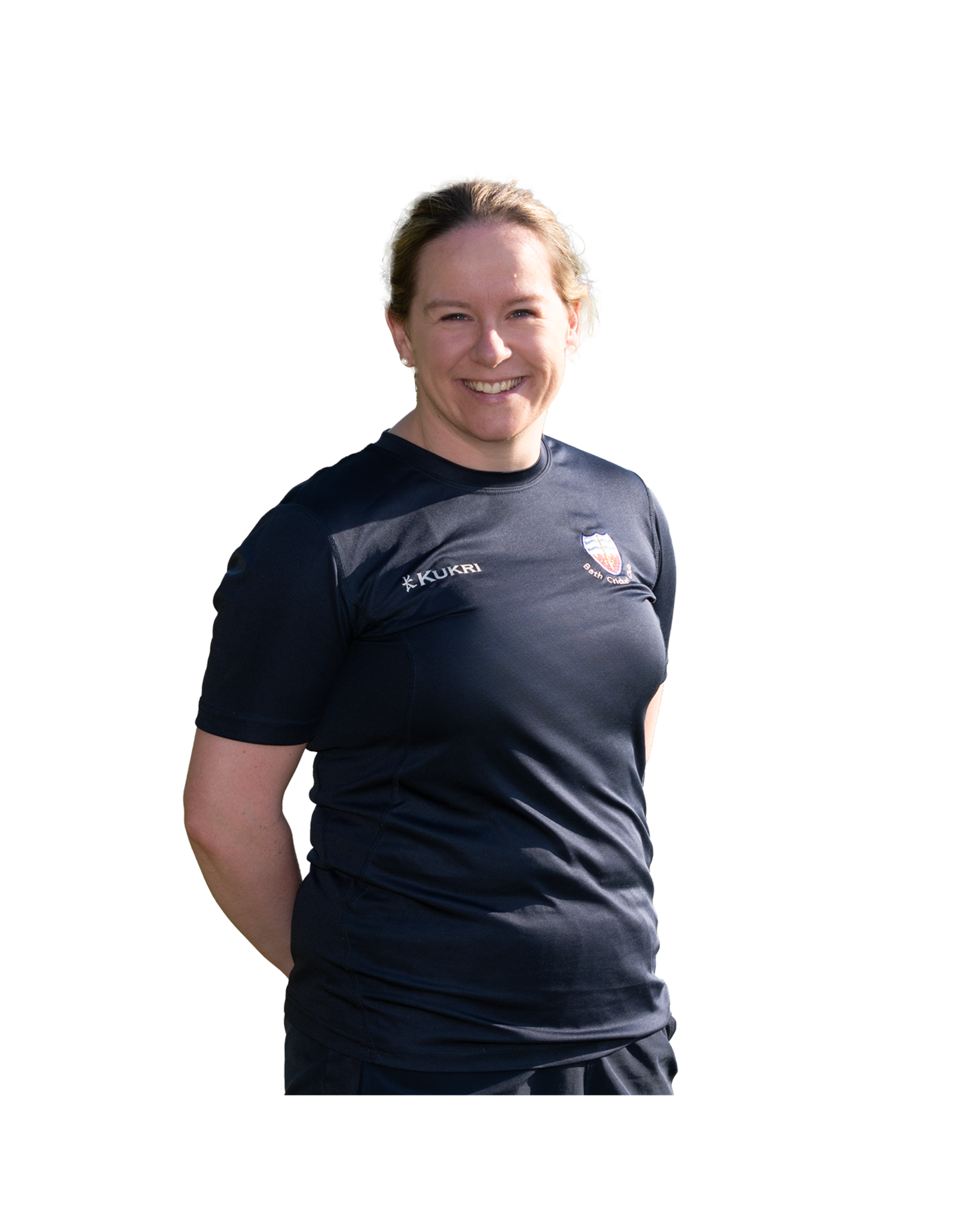 A woman with short blonde hair smiling, wearing a black sports shirt with logos that include 'Kukri' and a crest of Bath Cricket Club, standing outdoors.