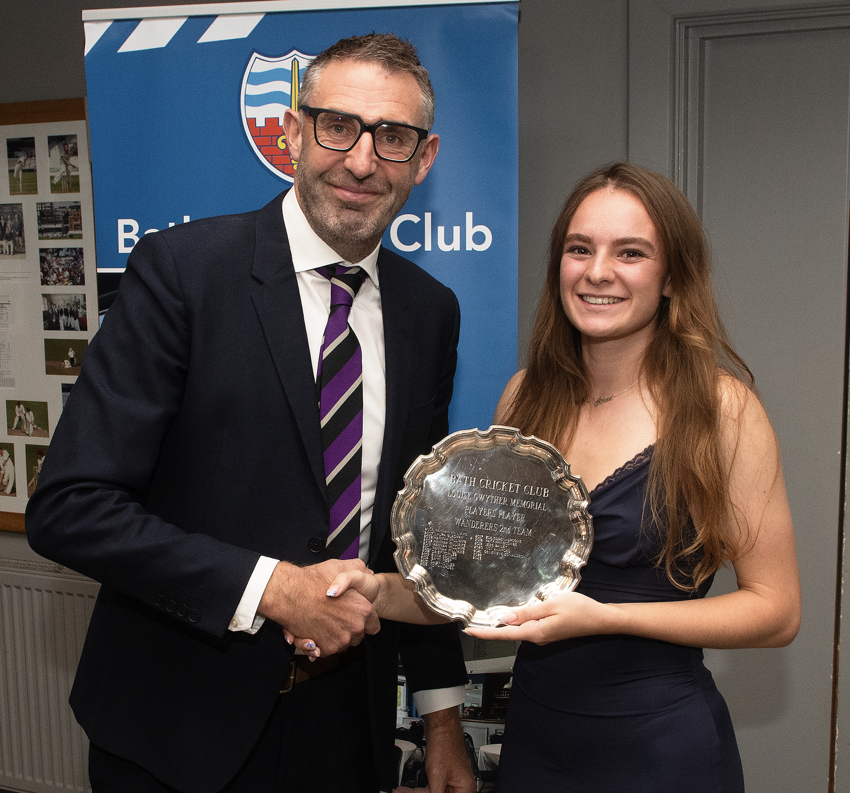A man in a suit with glasses shaking hands with a woman in a navy blue dress. They are holding an award plaque together, and both are smiling. In the background, there is a blue banner with a crest and the words 'Bath Cricket Club.'
