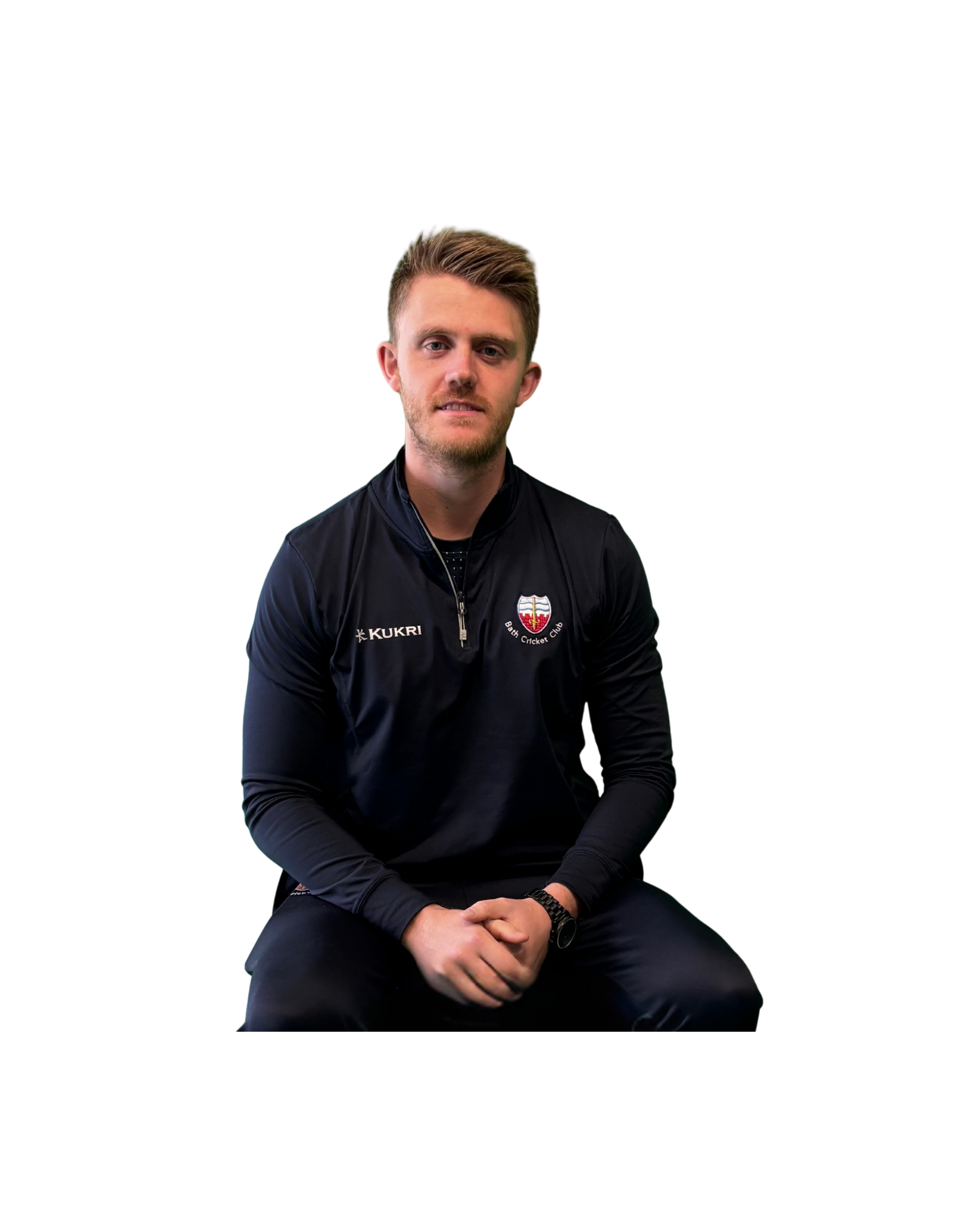 A young man dressed in a black jacket with a logo that says 'BATH CRICKET CLUB' on it, sitting with his hands clasped in front of him, against a plain black background.