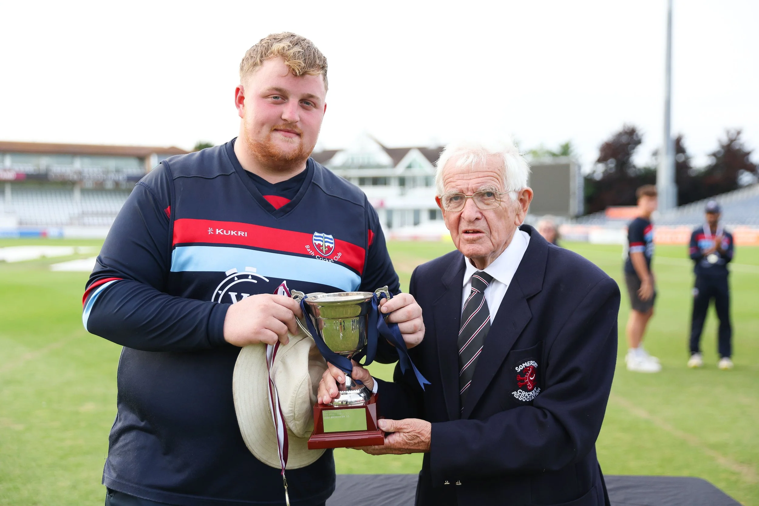 A young man in a sports jersey is holding a trophy, standing beside an older man in a suit and tie who is also holding the trophy. They are on a sports field, likely celebrating a victory.