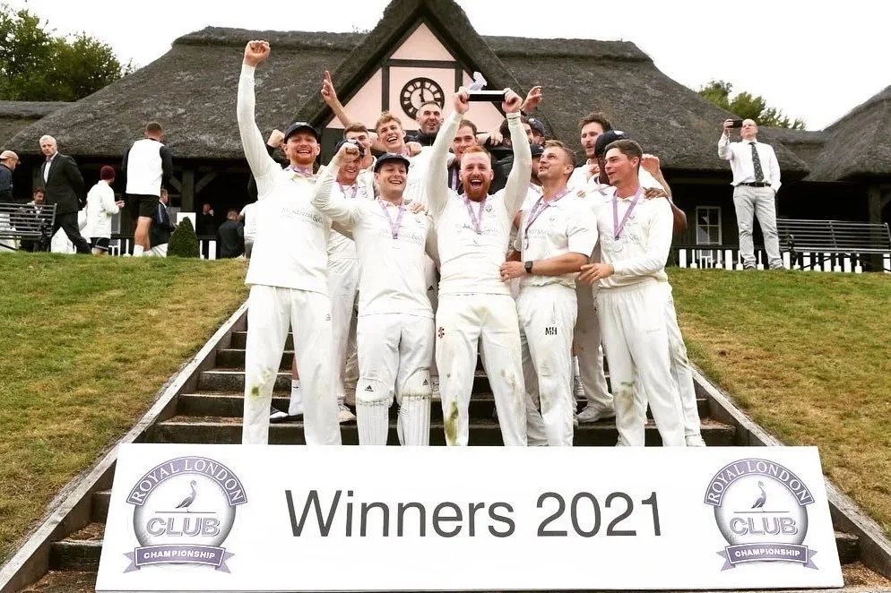 A group of cricket players celebrating as winners of the 2021 Royal London Club Championship, standing on stairs in front of a house with a thatched roof, holding a trophy and smiling.