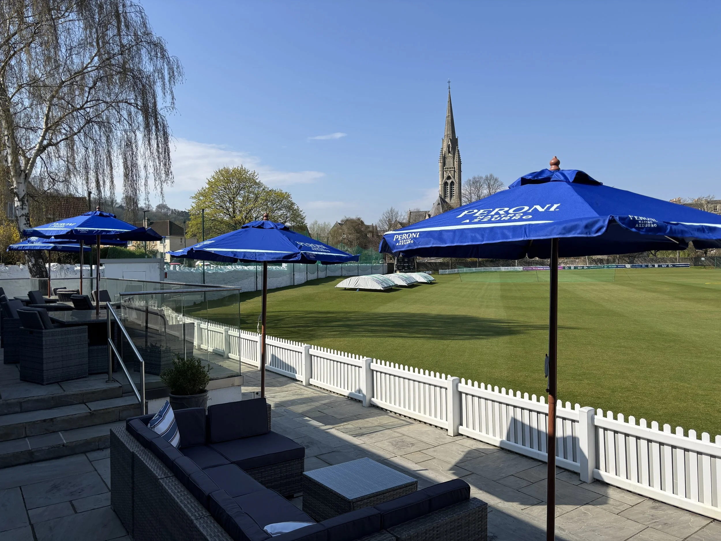Outdoor patio with wicker furniture and blue umbrellas overlooking a green sports field, church steeple, trees, and a clear blue sky.