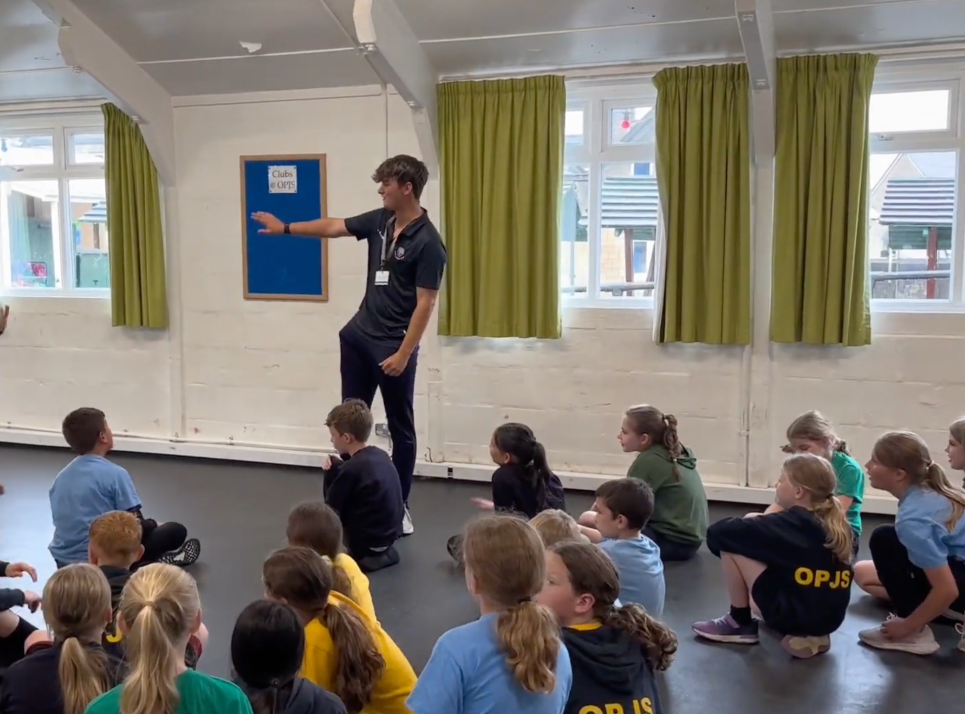 A classroom with students sitting on the floor, listening to a young male teacher standing in front of the class and gesturing with his arm. The room has large windows with green curtains and a blue bulletin board.