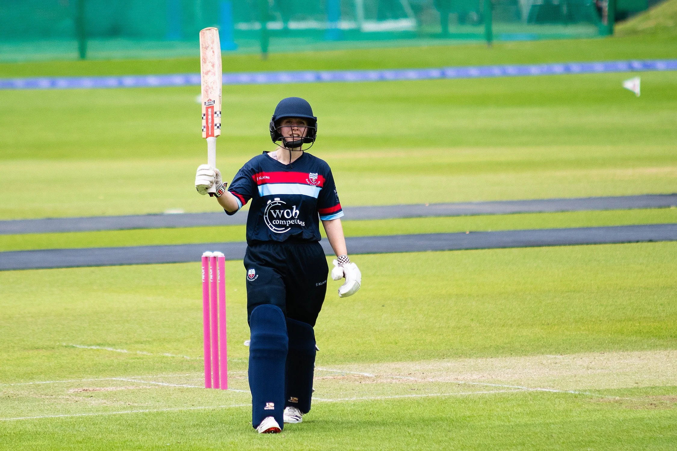 A young cricket player in a navy blue uniform with red, white, and blue stripes, wearing a helmet and gloves, standing on a cricket field near the stumps, holding a cricket bat in one hand, with a green grassy field and fencing in the background.