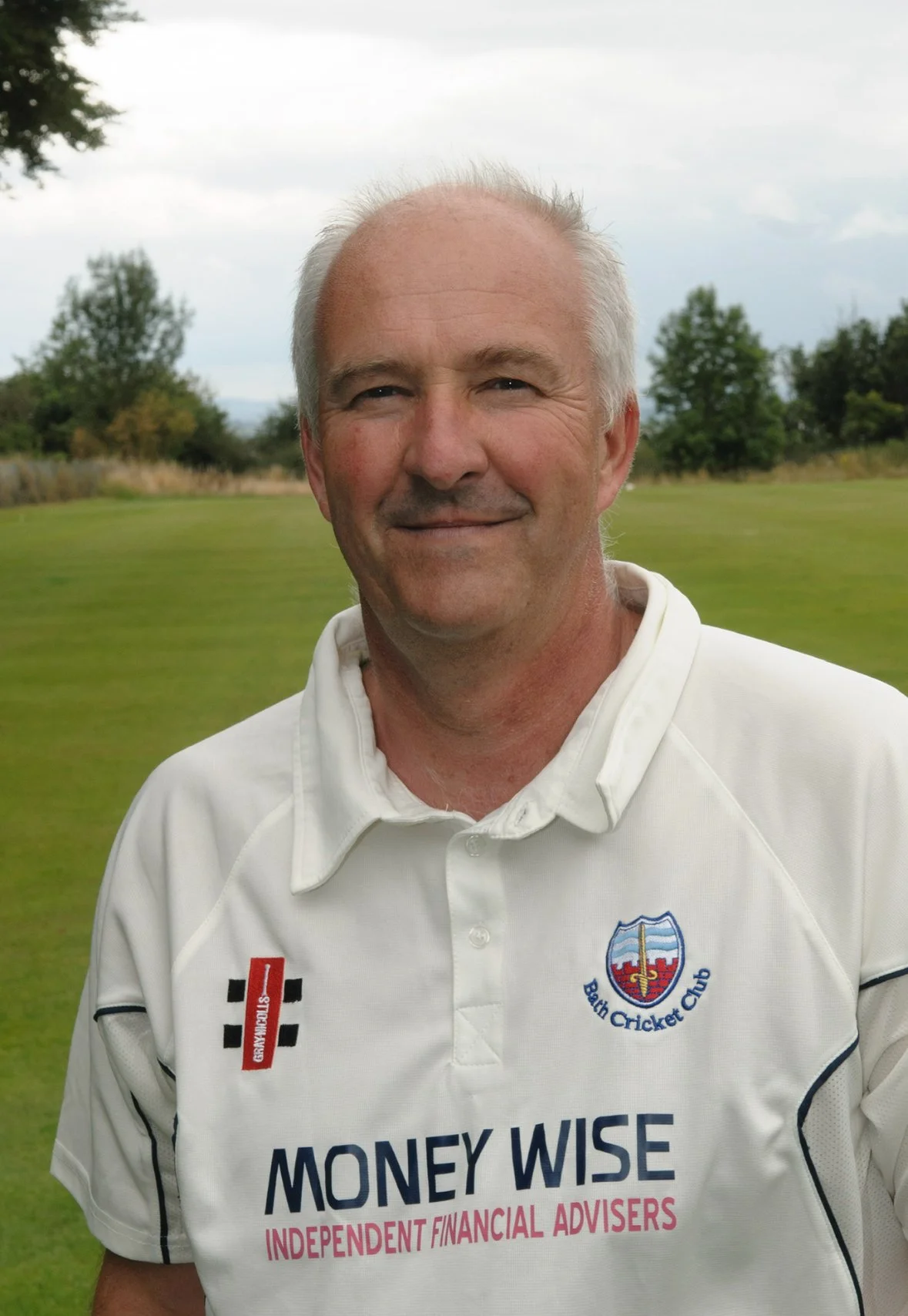 A middle-aged man with short gray hair smiling, standing outdoors on a grassy field with trees in the background. He is wearing a white sports shirt with logos, including 'Money Wise Independent Financial Advisers' and the Bath Cricket Club emblem.