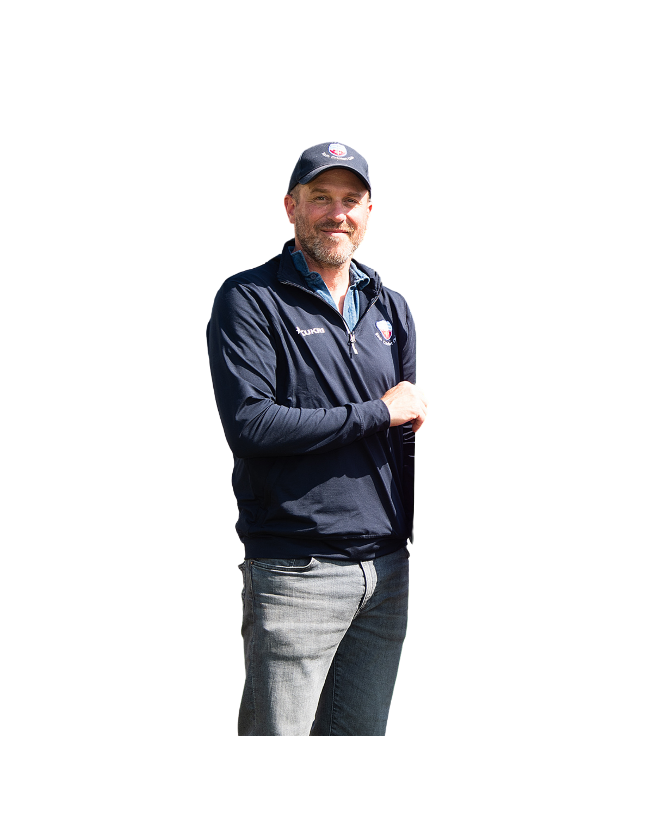 A man with a beard, wearing a black cap and a dark zip-up jacket with logos, standing with arms crossed, smiling outdoors.