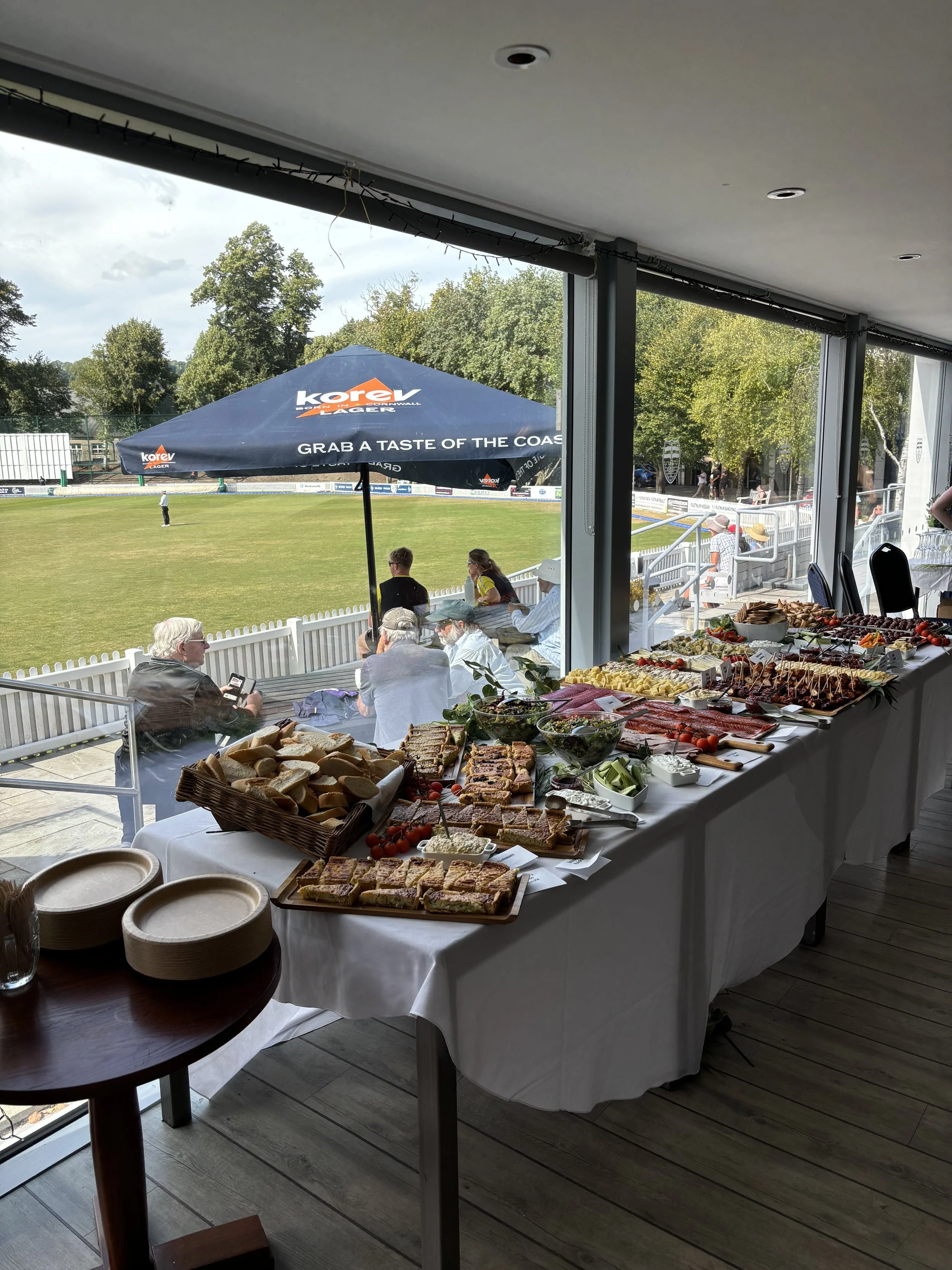 A buffet table with various finger foods including sandwiches, cheese, sliced meats, and fruit, set near large windows overlooking a grassy sports field. Outside, there are people sitting and standing, watching an event, with a large umbrella adverti