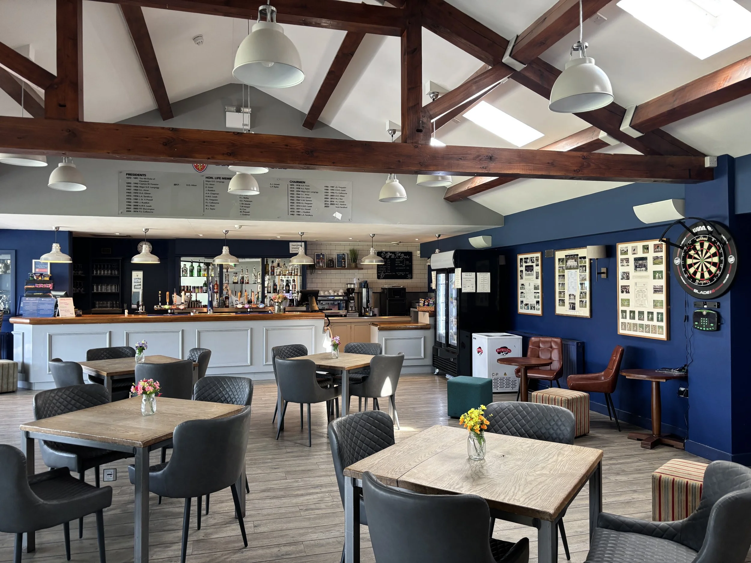 Interior of a café or restaurant with wooden beams on the ceiling, pendant lights, and tables with small flower arrangements, a bar area with bottles, and framed pictures on the walls.