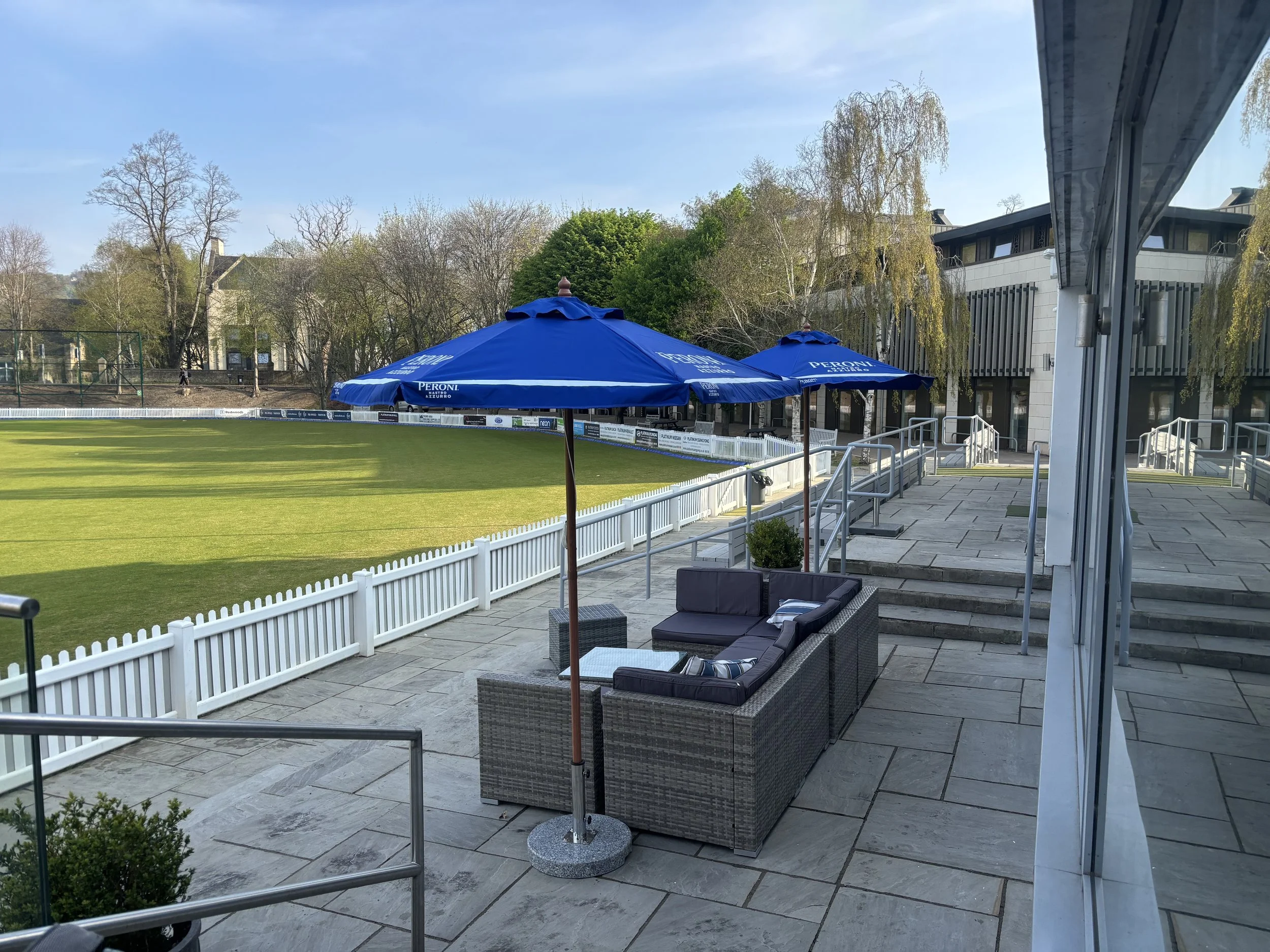 View of a tennis field with green grass, surrounded by a white fence, with blue umbrellas labeled Peroni, and outdoor seating with black cushions on a patio. There are trees and buildings in the background under a clear sky.